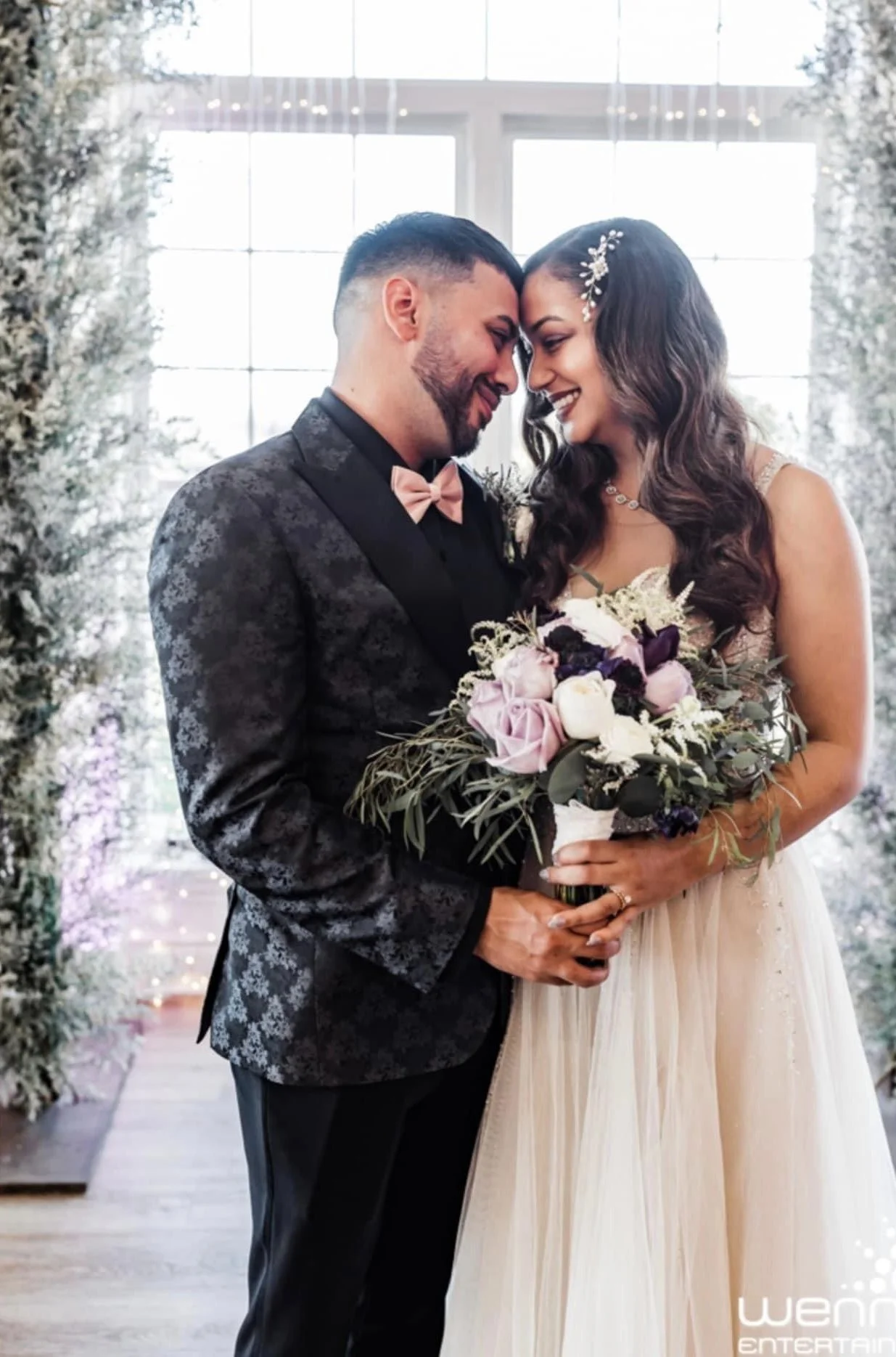 A bride and groom are smiling and touching foreheads, holding a bouquet of flowers, in a bright, floral setting.