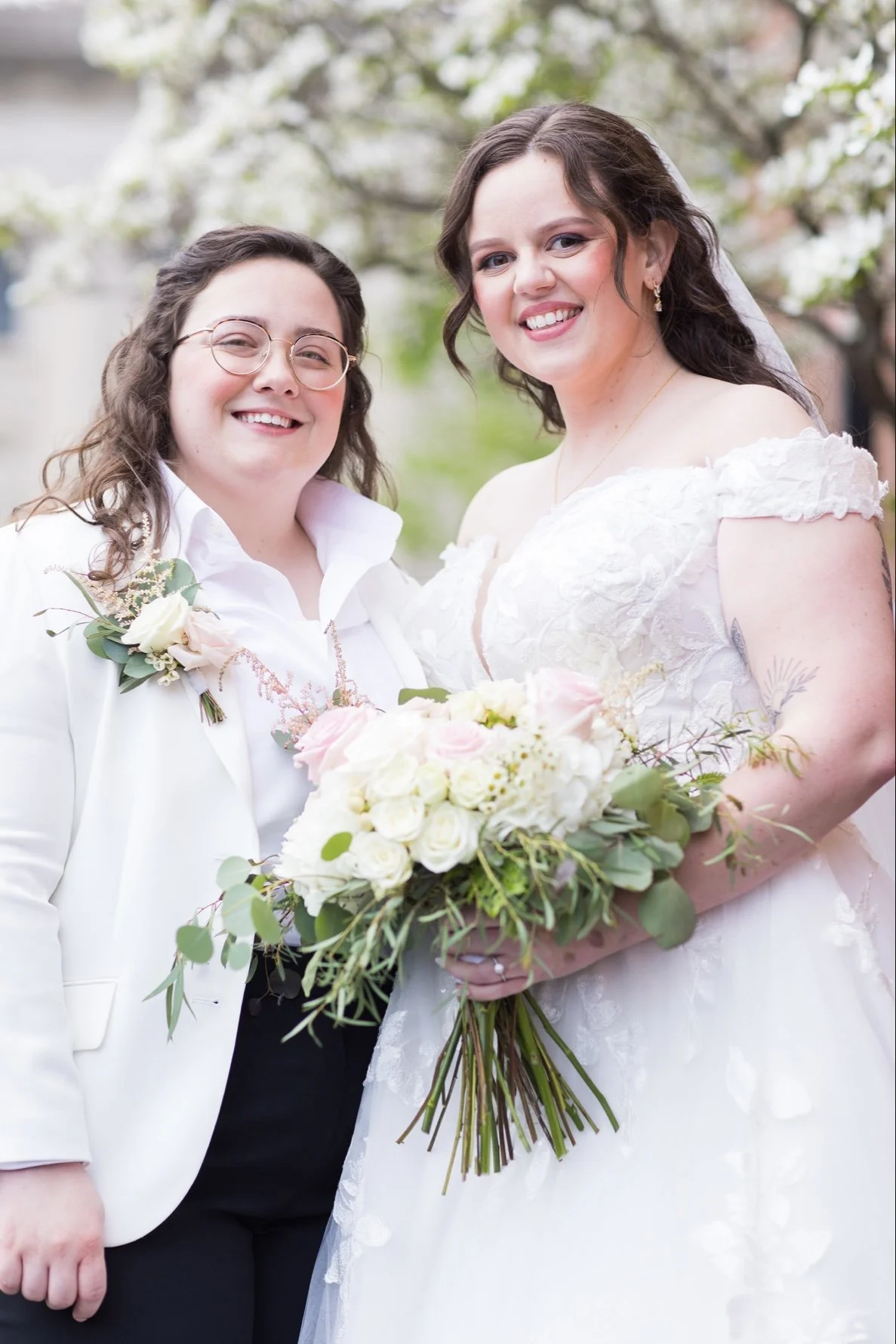 Two women, one in a white tuxedo and the other in a white wedding dress, standing outdoors, smiling, with flowers and greenery in the background.