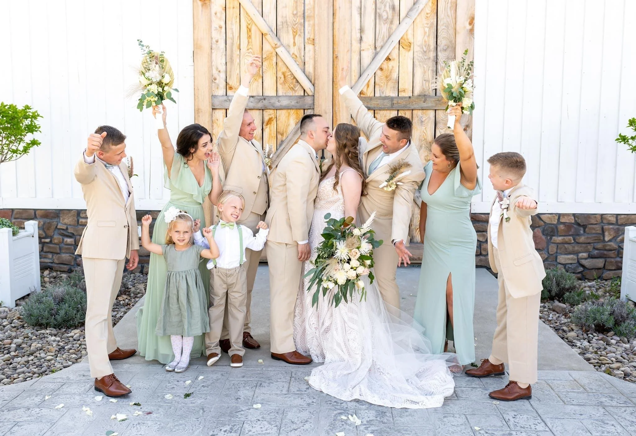 A wedding celebration with a bride and groom kissing, surrounded by family and friends in front of a rustic wooden gate. The group is celebrating joyfully with some holding bouquets, and a few children are flexing their arms in excitement.