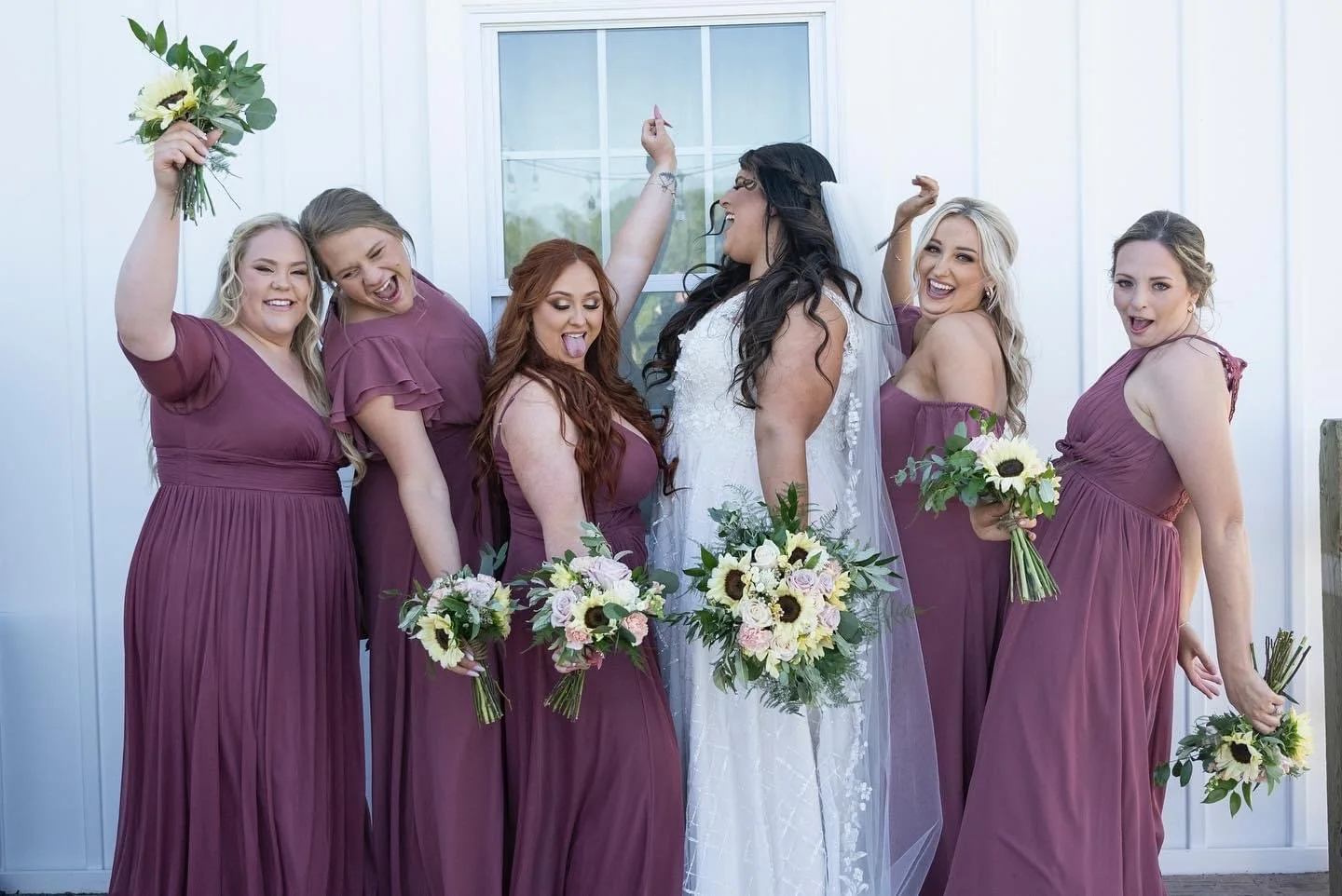 A bride in a white wedding dress surrounded by six bridesmaids in matching mauve dresses, all holding bouquets of flowers, celebrating together outside a building.