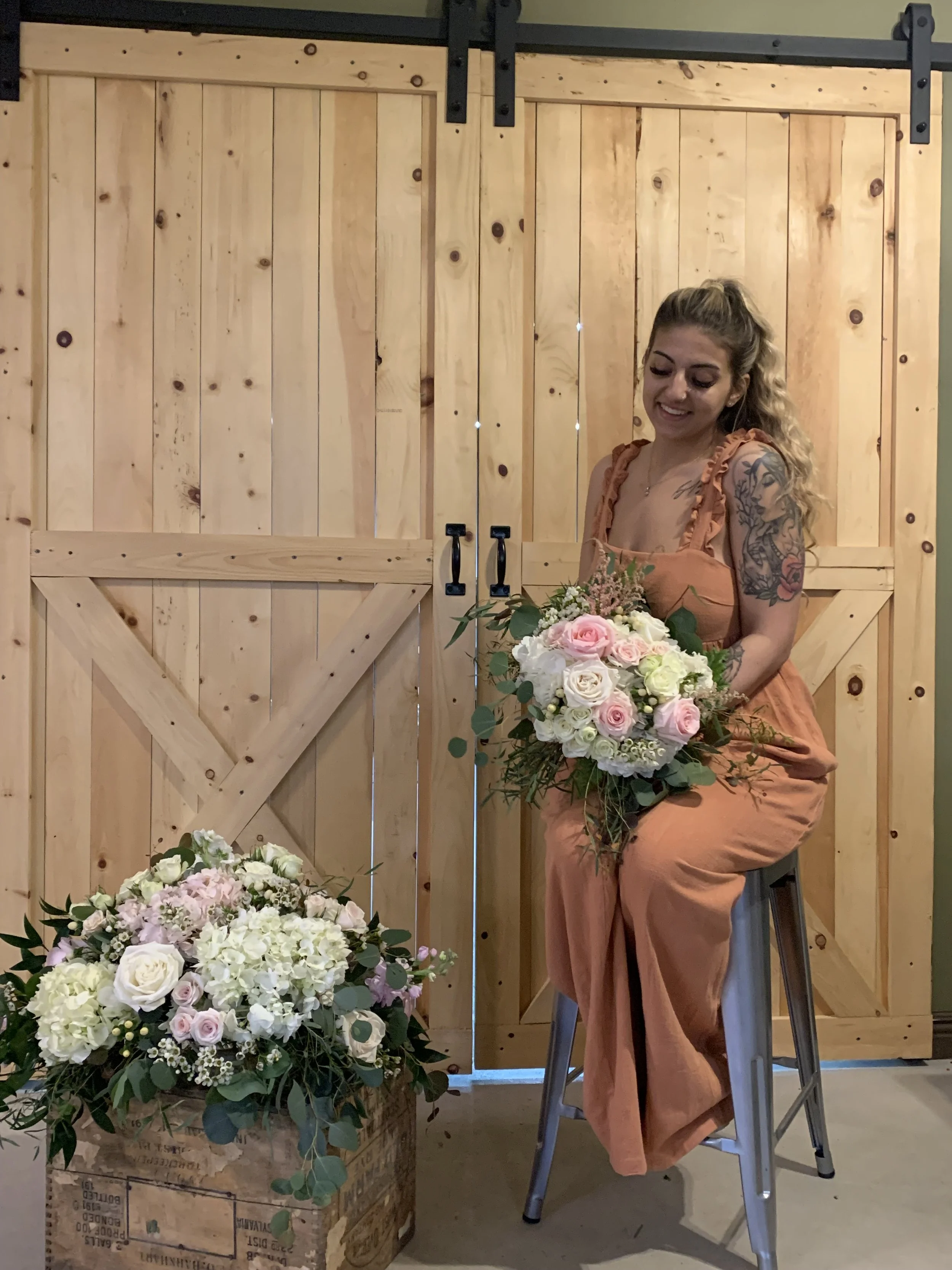A woman with tattoos on her arm, wearing an orange jumpsuit, sitting on a stool holding a large bouquet of white and pink roses, in front of wooden barn doors, with a smaller bouquet on the floor next to her.