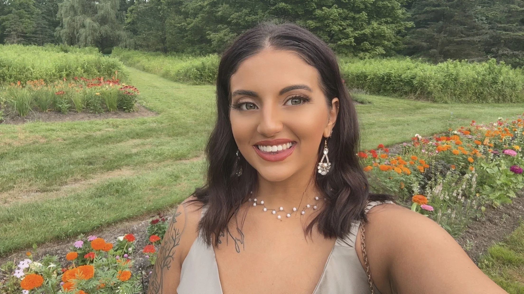 Young woman with dark hair and earrings taking a selfie in a garden with colorful flowers and green trees in the background.