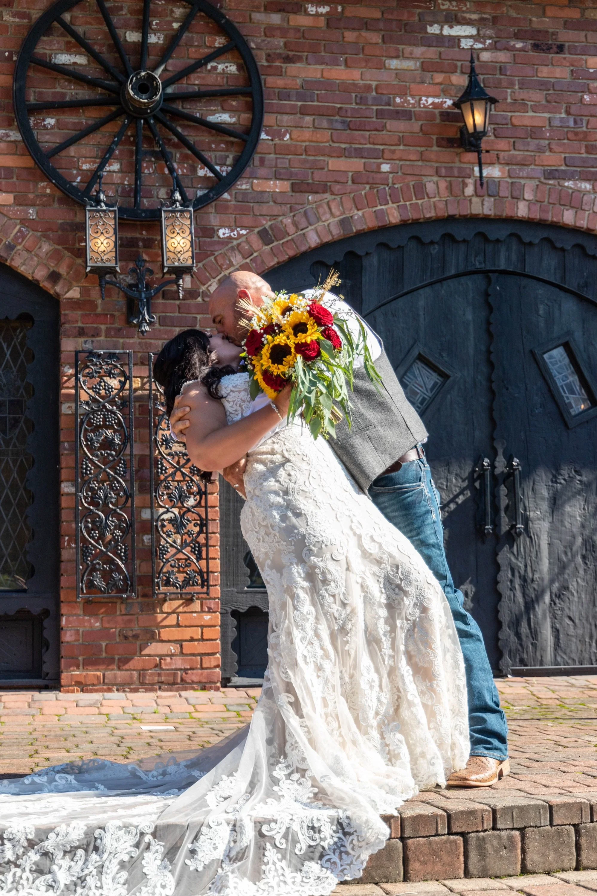 A bride and groom share a kiss outdoors in front of a brick building, with the groom holding a bouquet of sunflowers and red flowers.