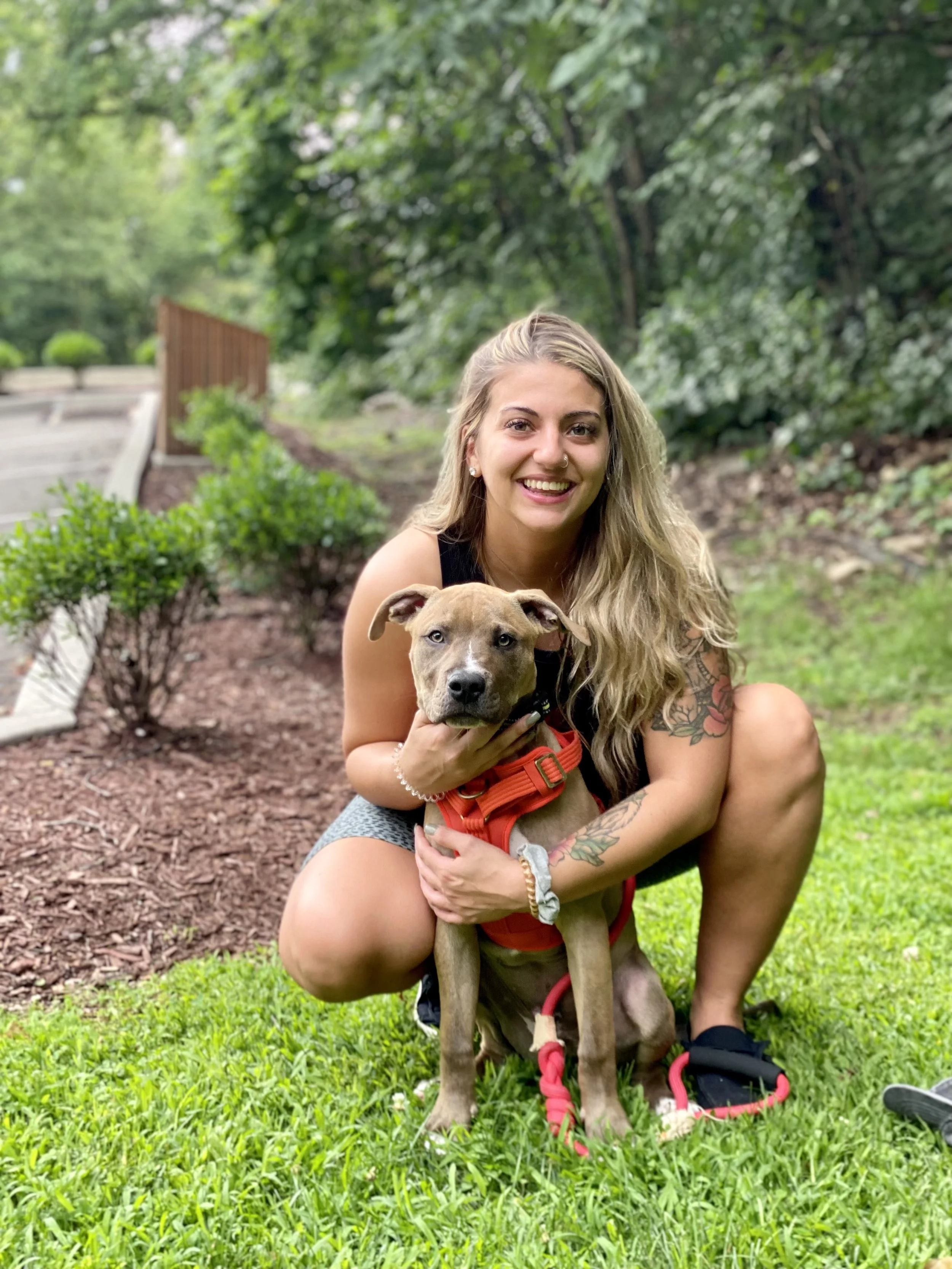 A young woman with long blonde hair, tattoos, and a black tank top crouches on the grass, smiling, while holding a brown dog with a red harness. They are outdoors in a park with green trees and shrubs in the background.