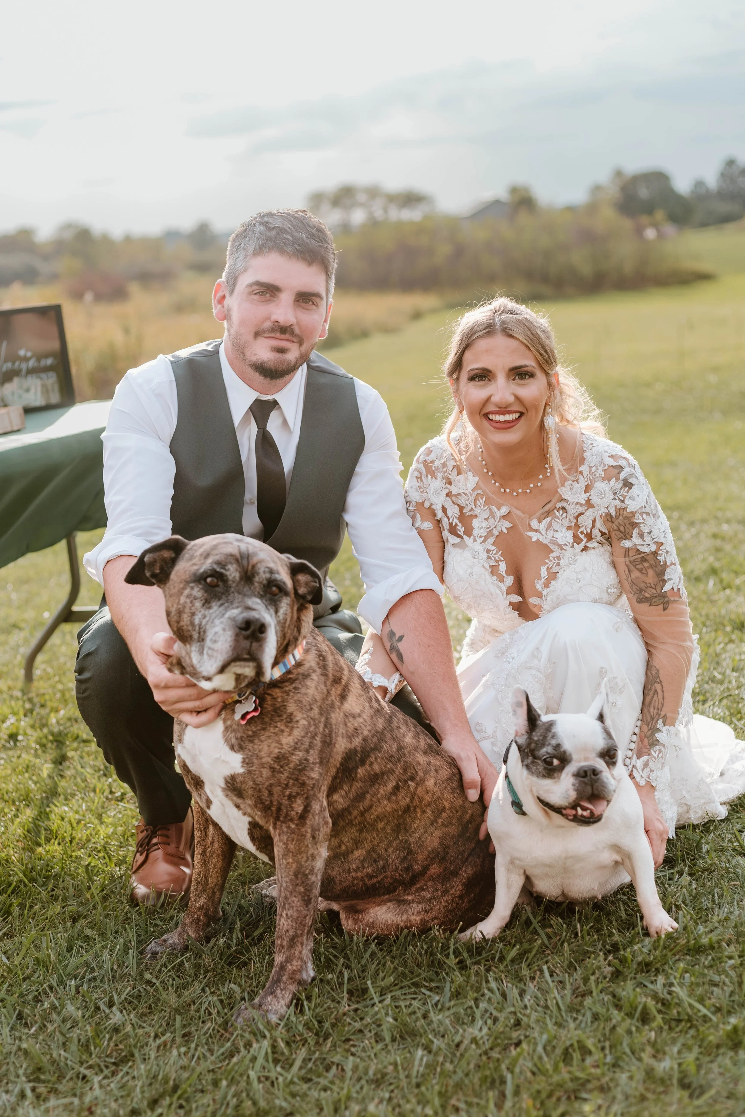 A newlywed couple posing outdoors on a grassy field with their two dogs, one large brindle and one small white with black spots, during daytime.