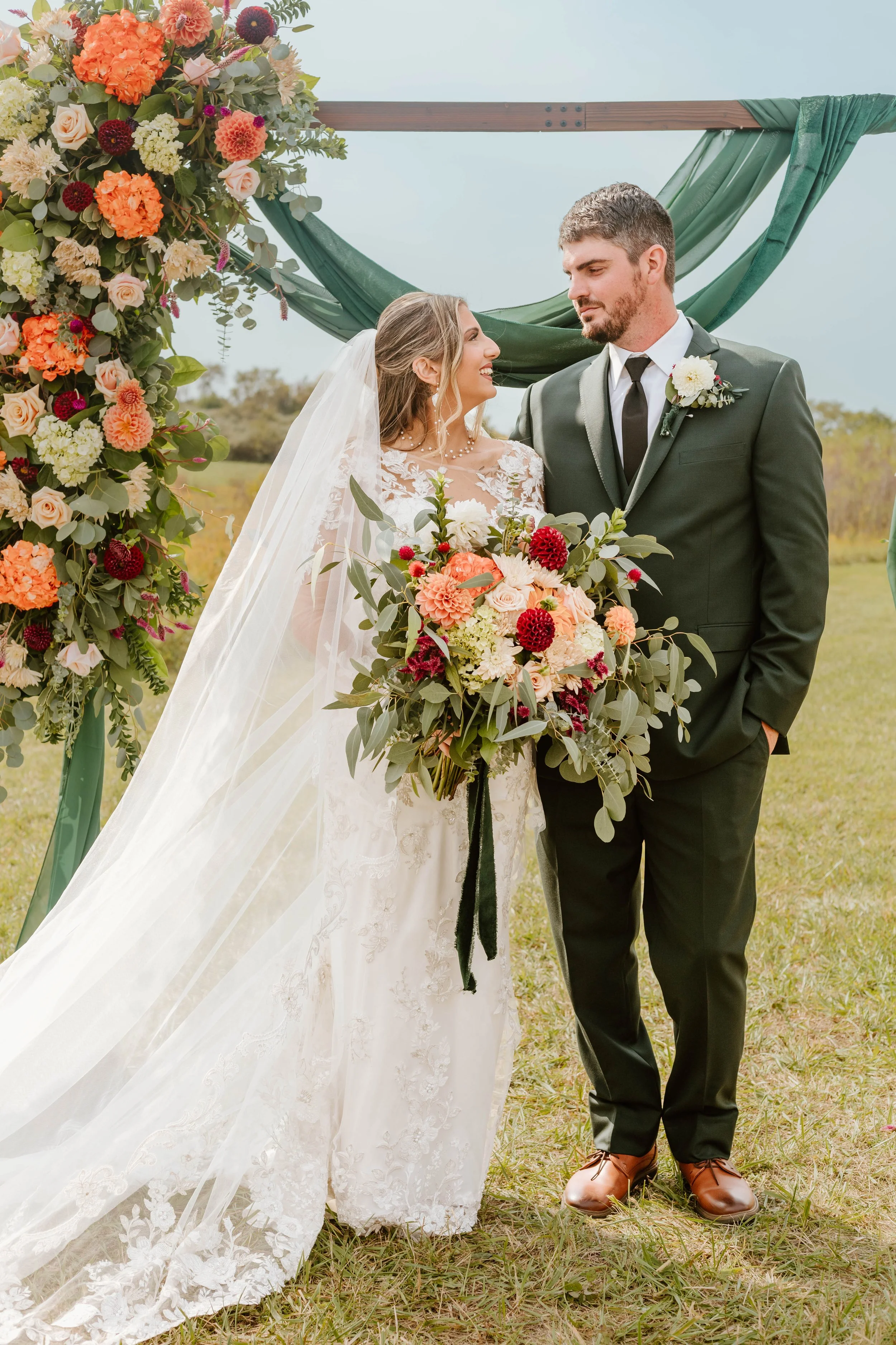 A bride and groom stand outdoors under a floral arch during their wedding ceremony. The bride is holding a large bouquet, and the groom is dressed in a dark suit with a white flower boutonniere. They look lovingly at each other on a grassy field with a cloudy sky above.