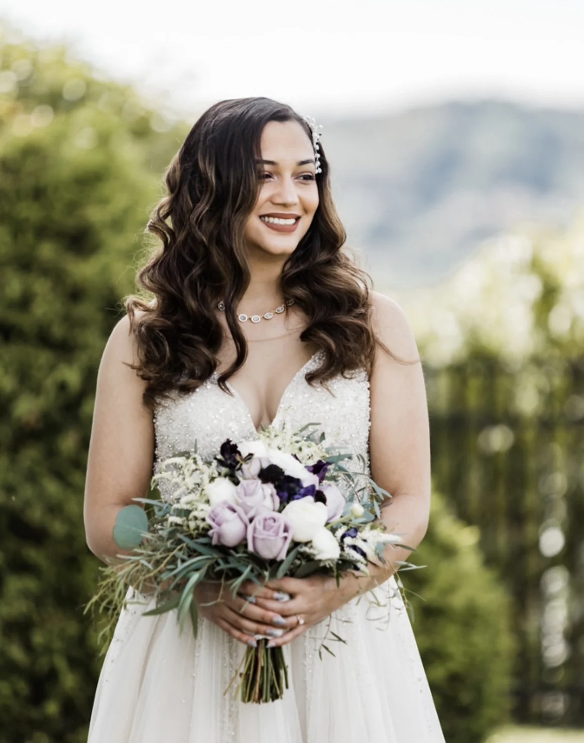 A bride in a white wedding dress holding a bouquet of purple and white flowers, standing outdoors with greenery in the background.