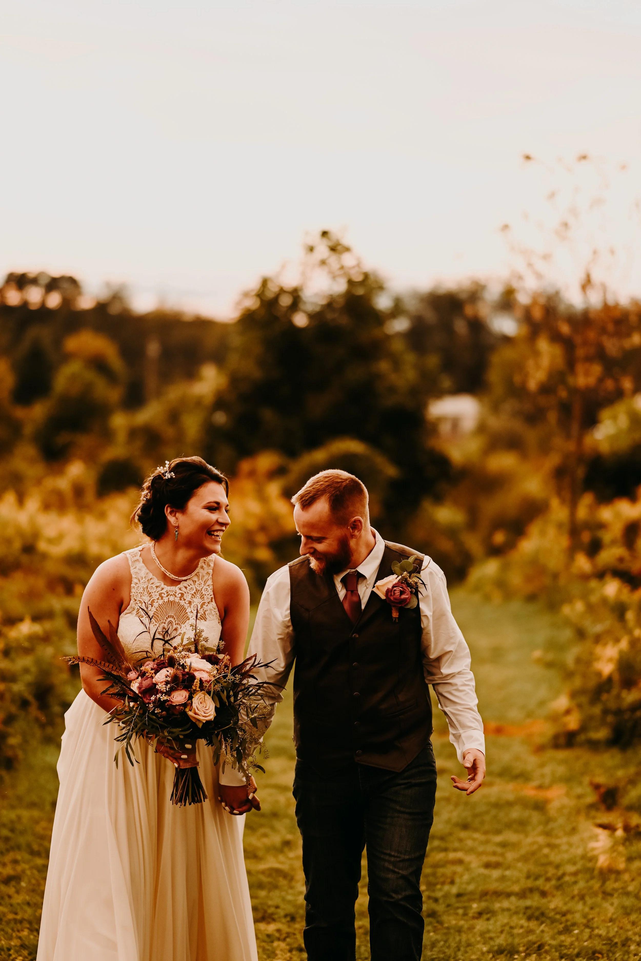A happy bride and groom walking outdoors during sunset, smiling and holding hands, with a scenic background of trees and sky.
