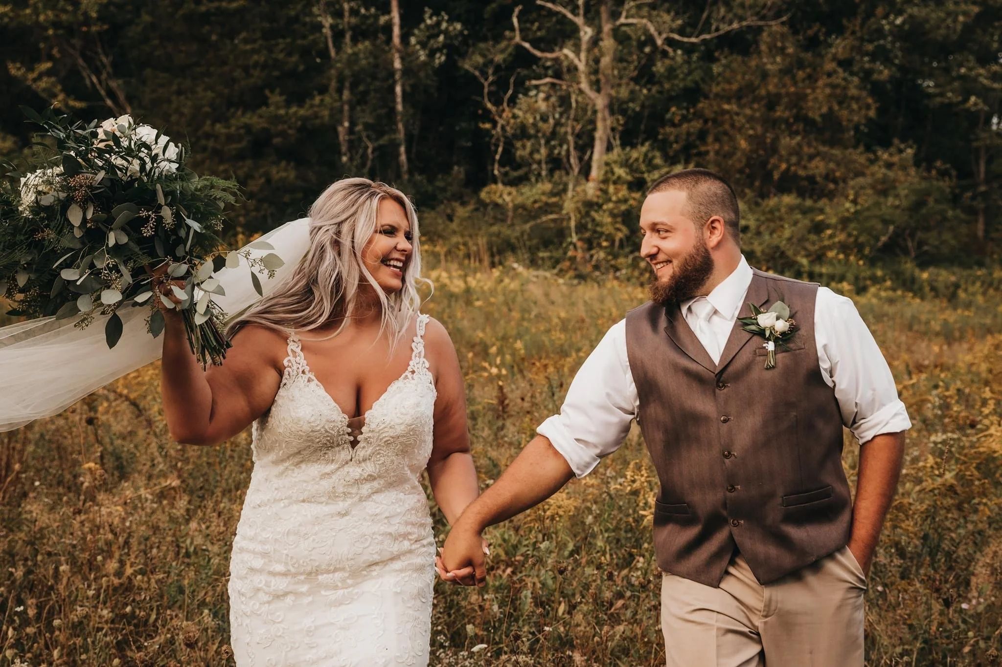 A smiling woman in a wedding dress holding a bouquet, and a man in a vest and rolled-up sleeves holding her hand, walking through a grassy field with trees in the background.