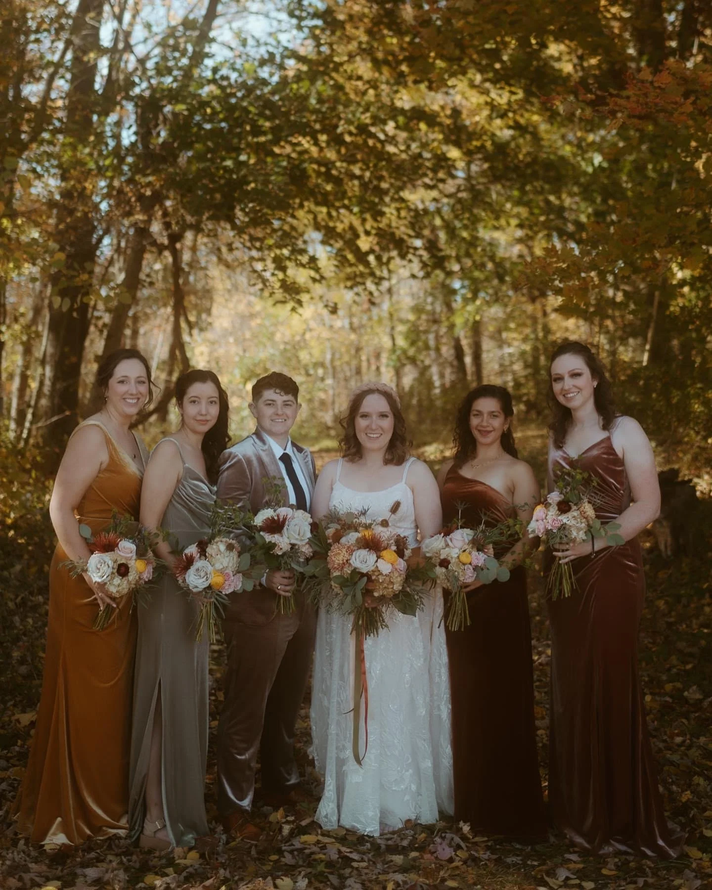Group of six women and one man in formal attire holding bouquets of flowers, standing outdoors on a fallen leaf-covered ground with trees and autumn leaves in the background.