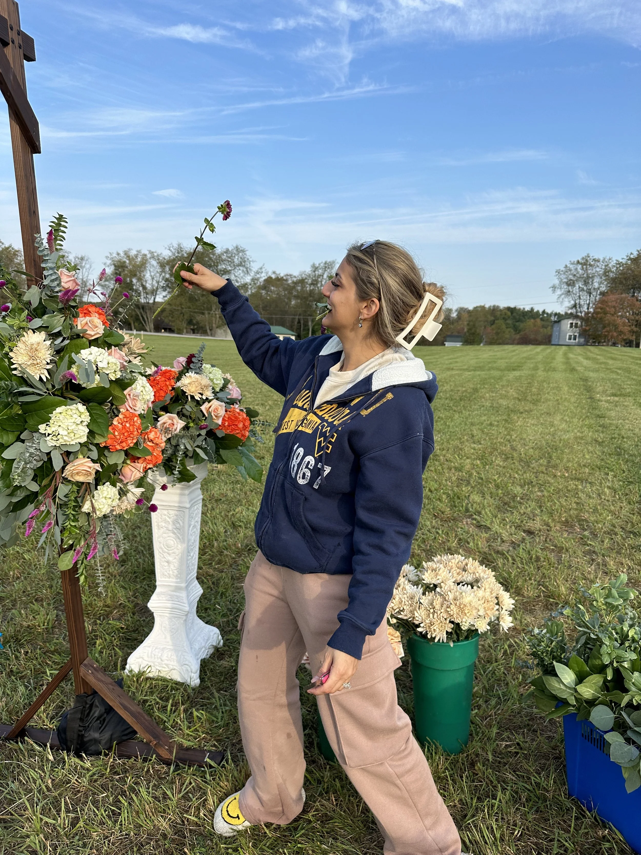 A woman in a navy hoodie and tan pants arranging colorful flowers on a floral display outdoors on a sunny day