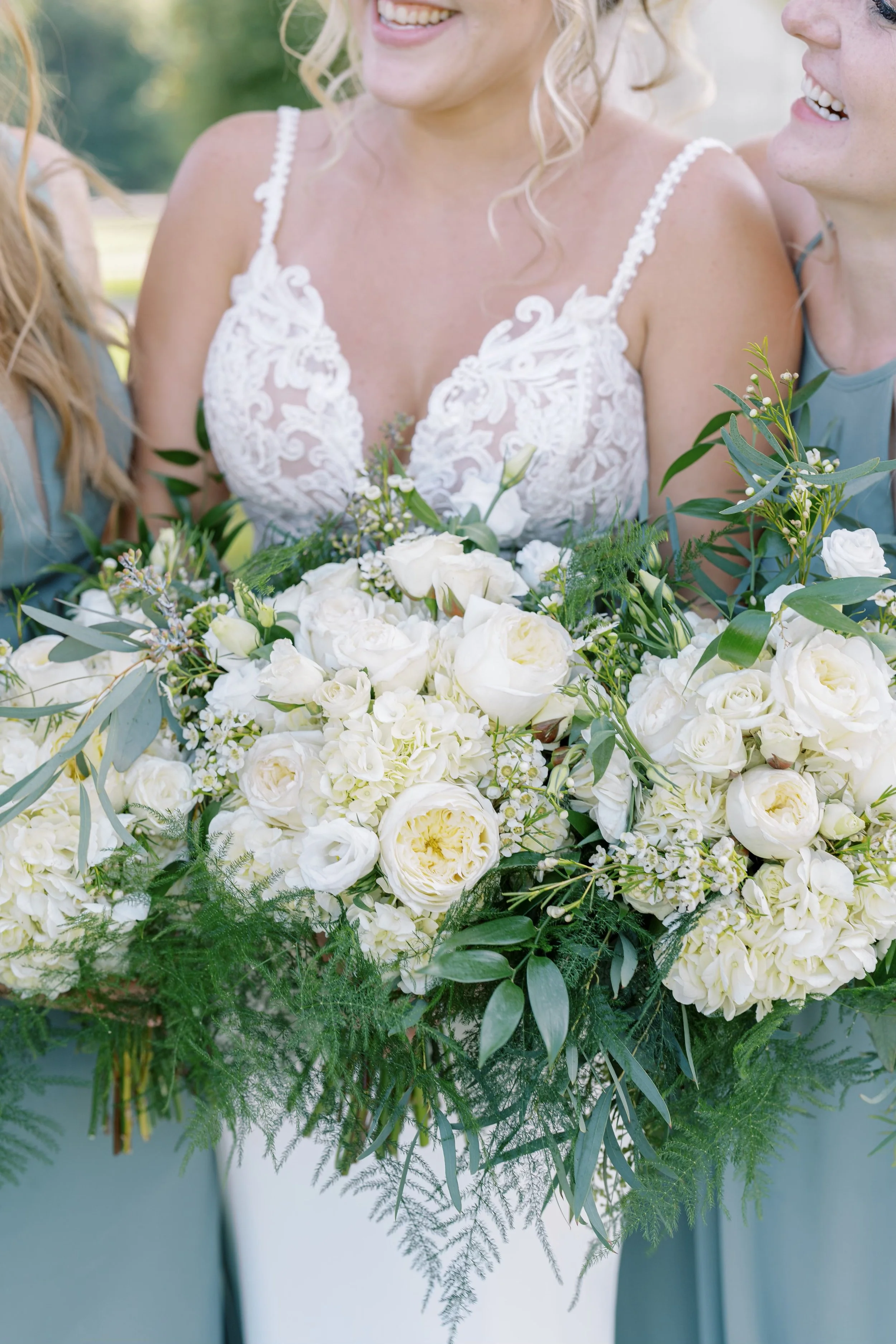 A bride in a white dress holding a large bouquet of white flowers and greenery, smiling with two women standing beside her, all outdoors on a sunny day.