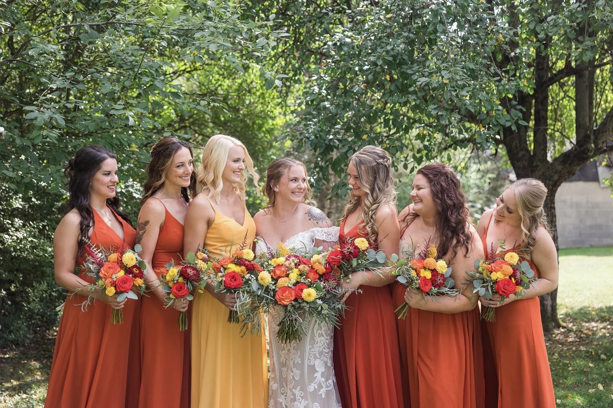 A bride and six bridesmaids standing outdoors on a sunny day, holding colorful bouquets, with green trees in the background.