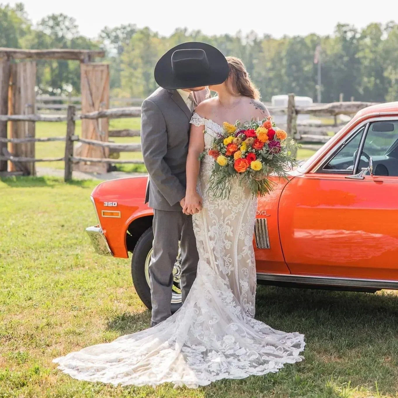 Bride and groom standing outdoors near a bright orange vintage car, kissing while holding hands, with the bride holding a large colorful bouquet, on a grassy field with a wooden fence and trees in the background.