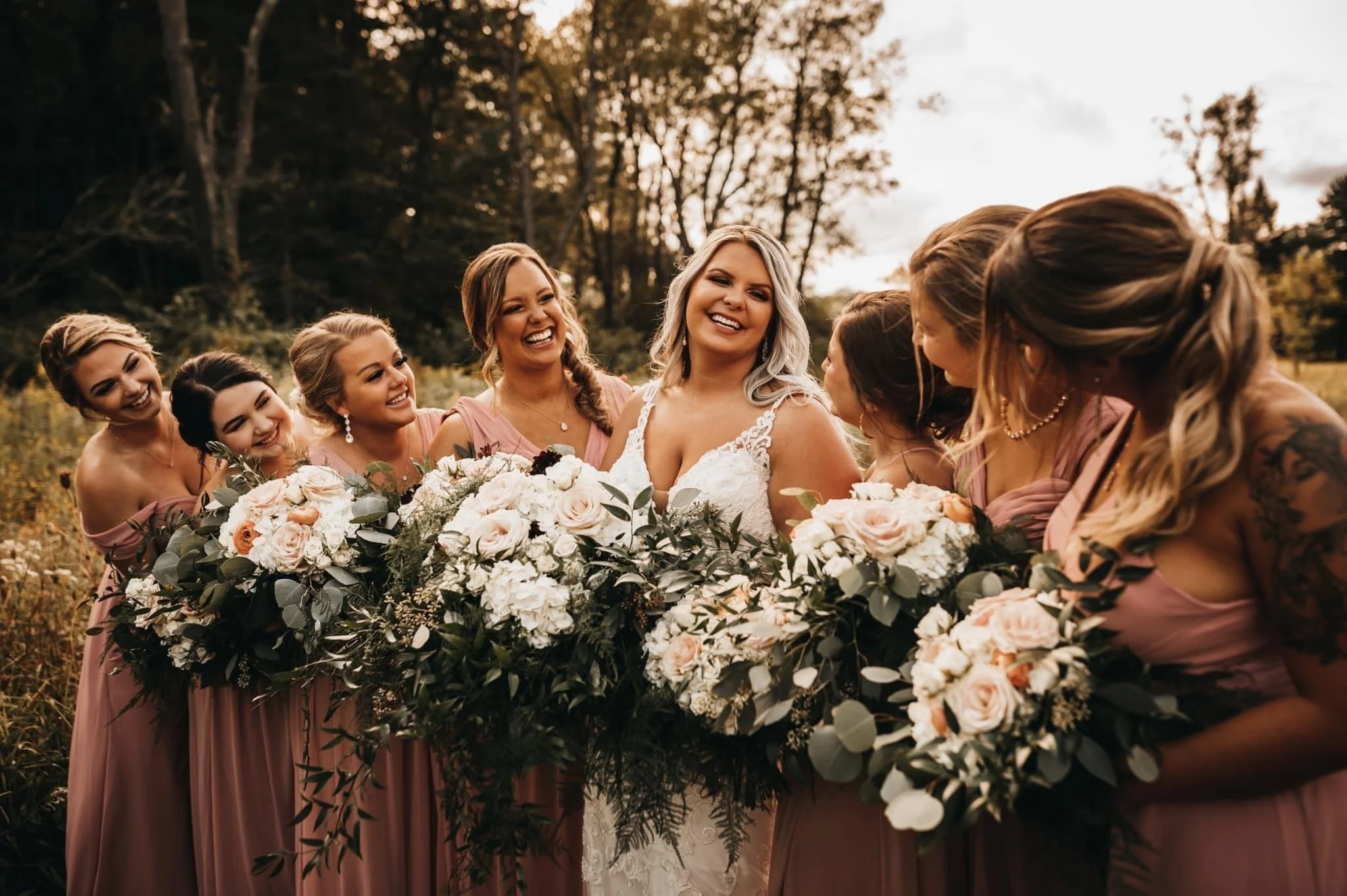 A bride with six bridesmaids smiling and holding large floral bouquets outdoors during daytime, in a natural setting with trees and a cloudy sky.
