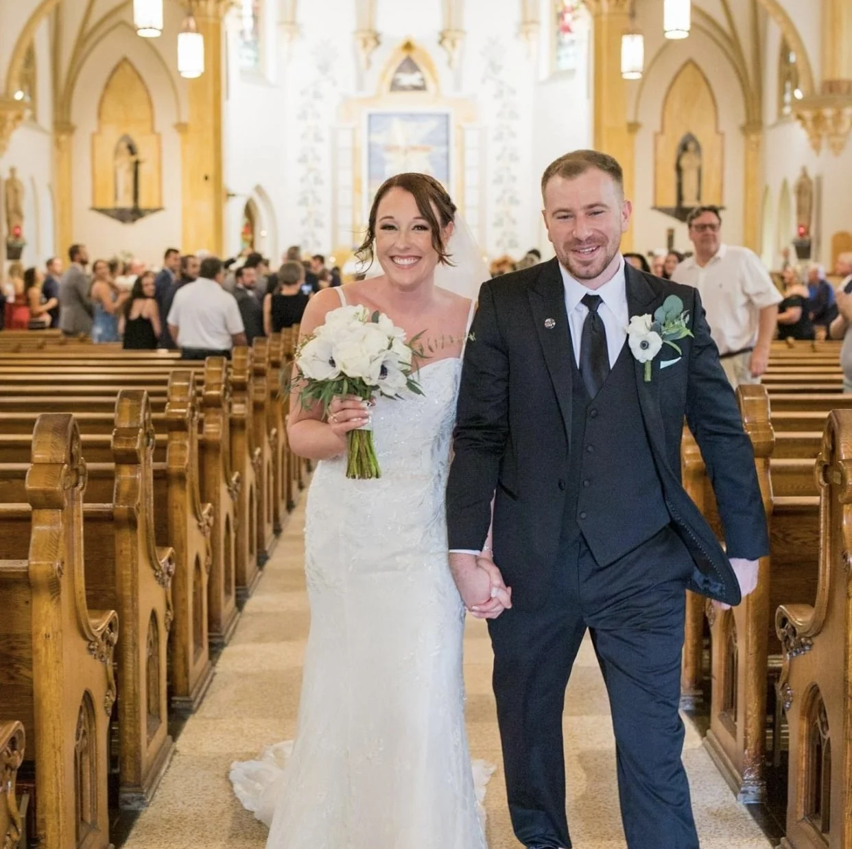 A bride and groom walking down the aisle holding hands in a church with wedding guests in the background.