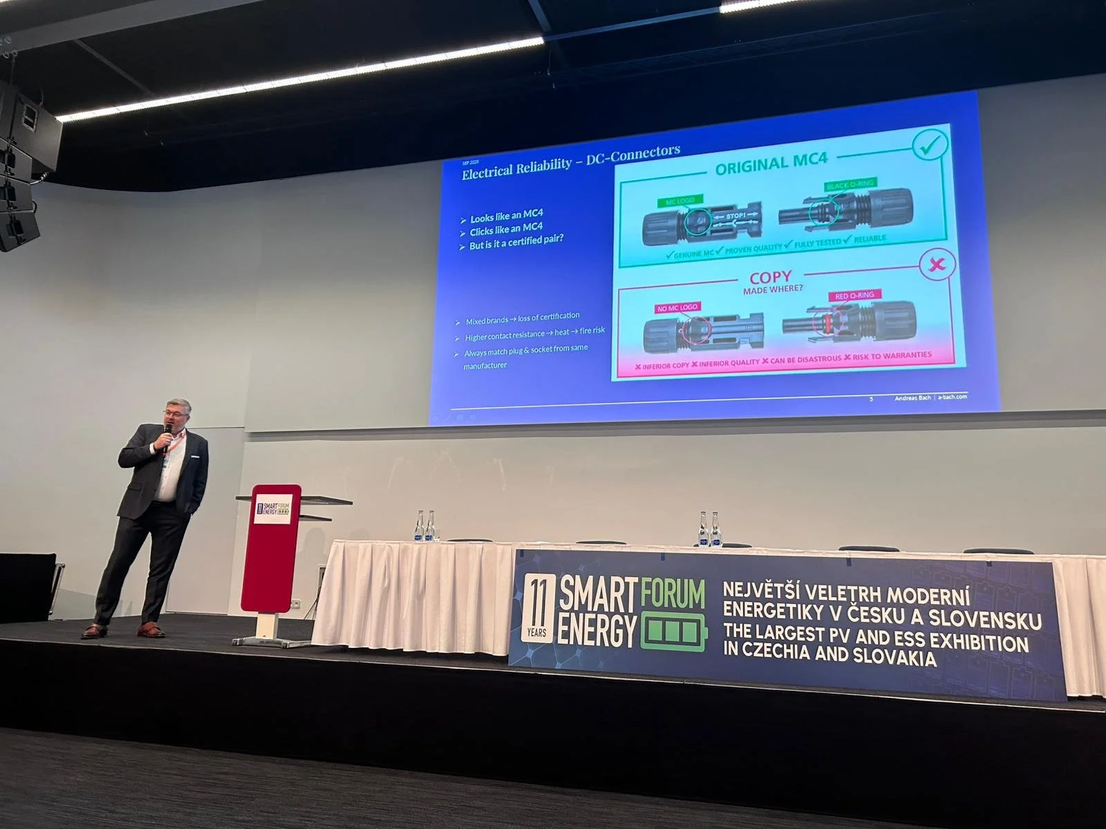 A man in a dark suit standing on a stage holding a microphone, giving a presentation. The large screen behind him displays slides about electrical reliability of DC connectors, comparing original and copied connectors. There is a long table with three water bottles and a sign in front of it, at the Smart Energy Forum in Czechia and Slovakia.