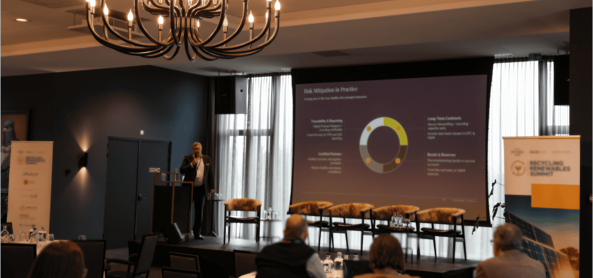 A conference room with a large screen displaying a presentation titled 'Risk Mitigation in Practice.' A man in a suit stands at a podium with a microphone, addressing the audience. Several chairs are on the stage, and some attendees are seated facing the stage. A decorative chandelier hangs from the ceiling, and sunlight filters through tall curtains.