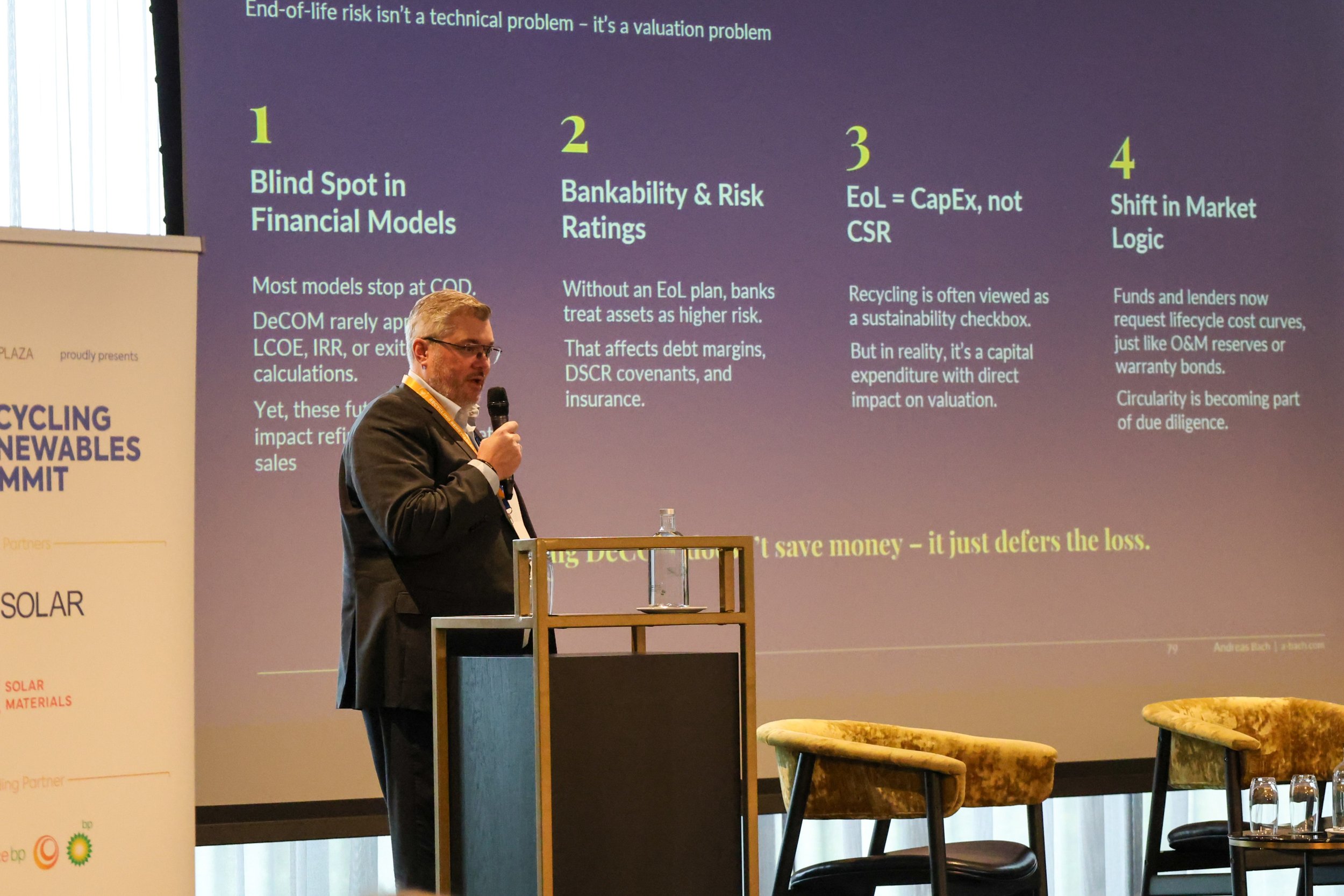 A man in a dark suit speaking into a microphone at a conference, standing next to a presentation slide titled 'End-of-life risk isn't a technical problem — it's a valuation problem'. The slide lists four points regarding financial and risk assessment concepts. There are three yellow chairs and a table with glasses on the stage in front of the man, and a vertical banner on the left side of the image.
