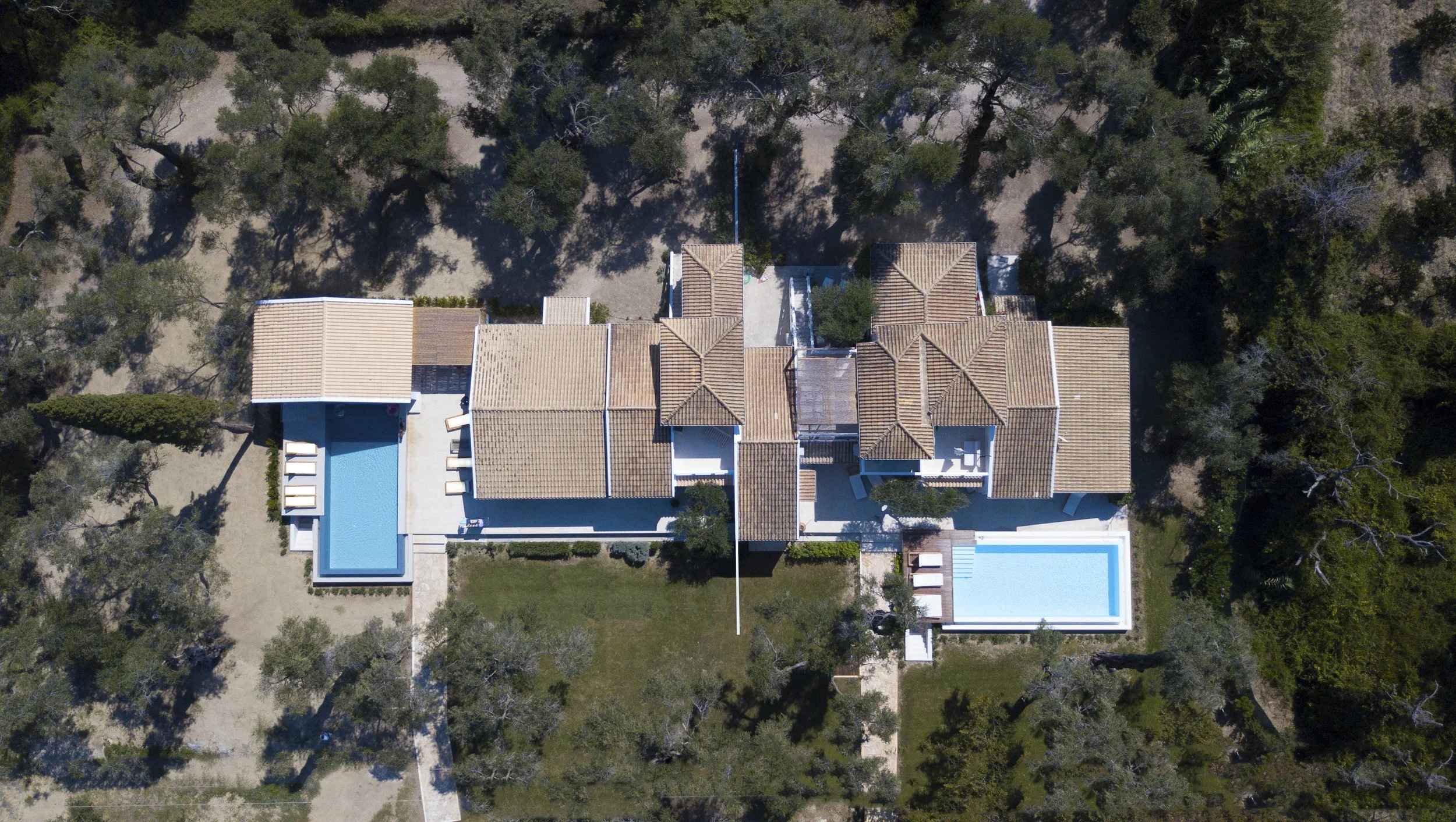 Aerial view of a large house with a tiled roof and two swimming pools, surrounded by trees with a dirt driveway leading to the property.