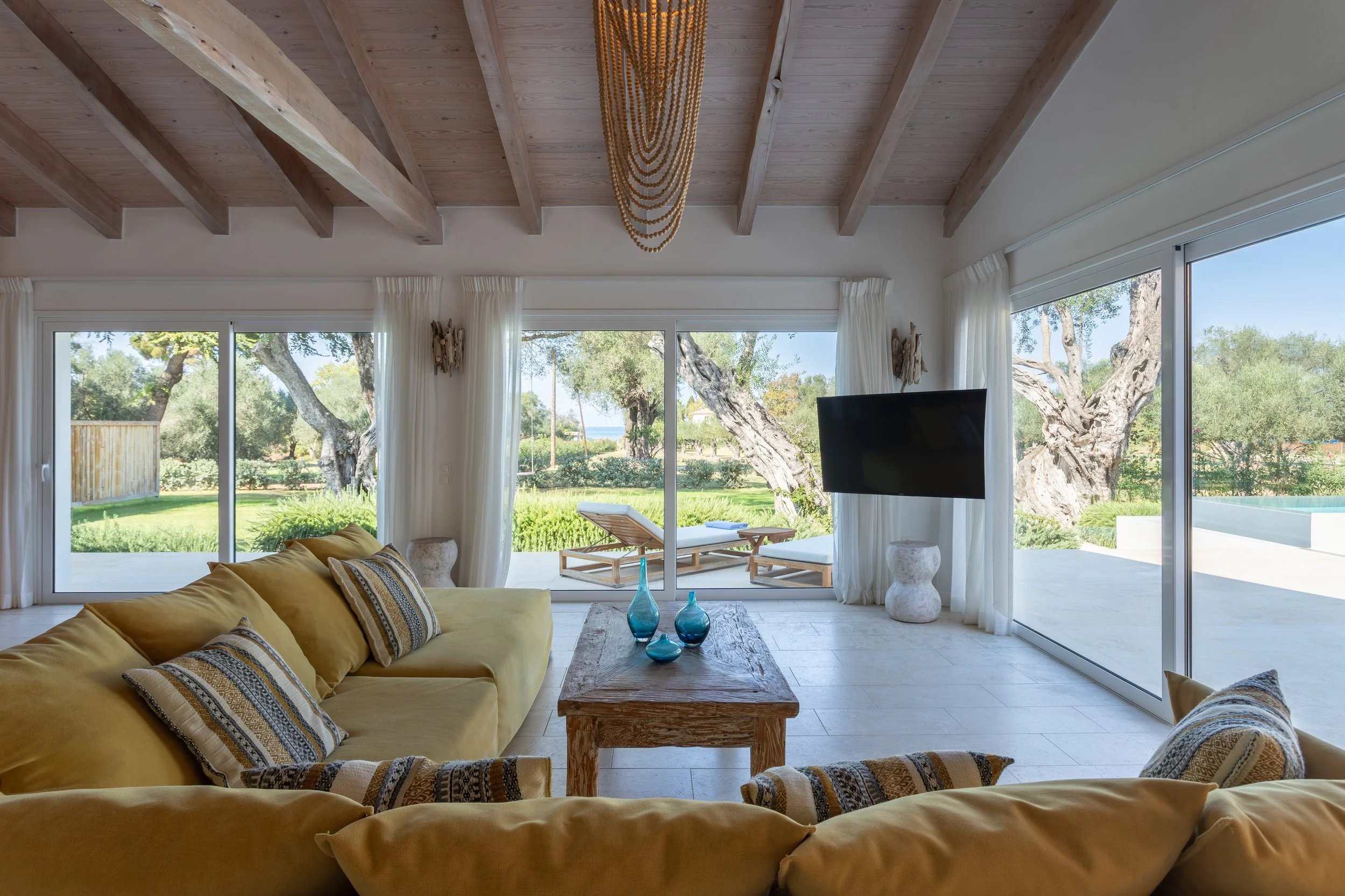 Living room with yellow sofas, a wooden coffee table with blue vases, large glass sliding doors, white curtains, and outdoor lounge chairs overlooking a green yard with trees.