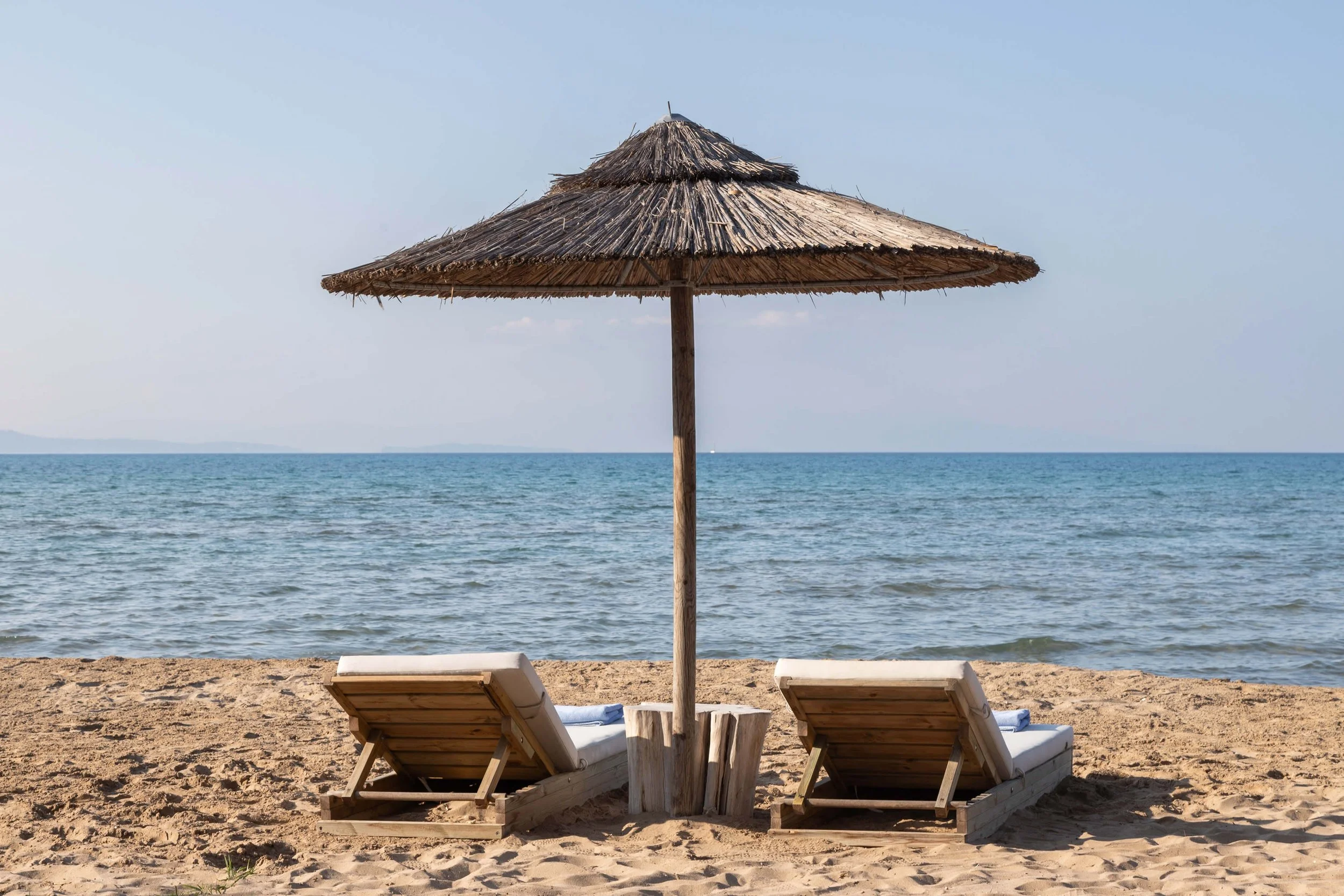 Beach scene with two wooden lounge chairs and a thatched umbrella on sandy shore, ocean in background, clear sky.