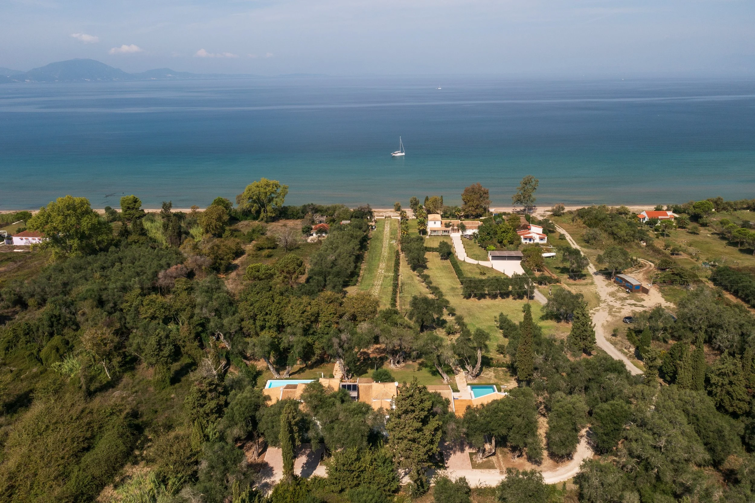 Aerial view of a coastal residential area with houses, trees, and swimming pools, overlooking a sandy beach and calm ocean with a sailboat.