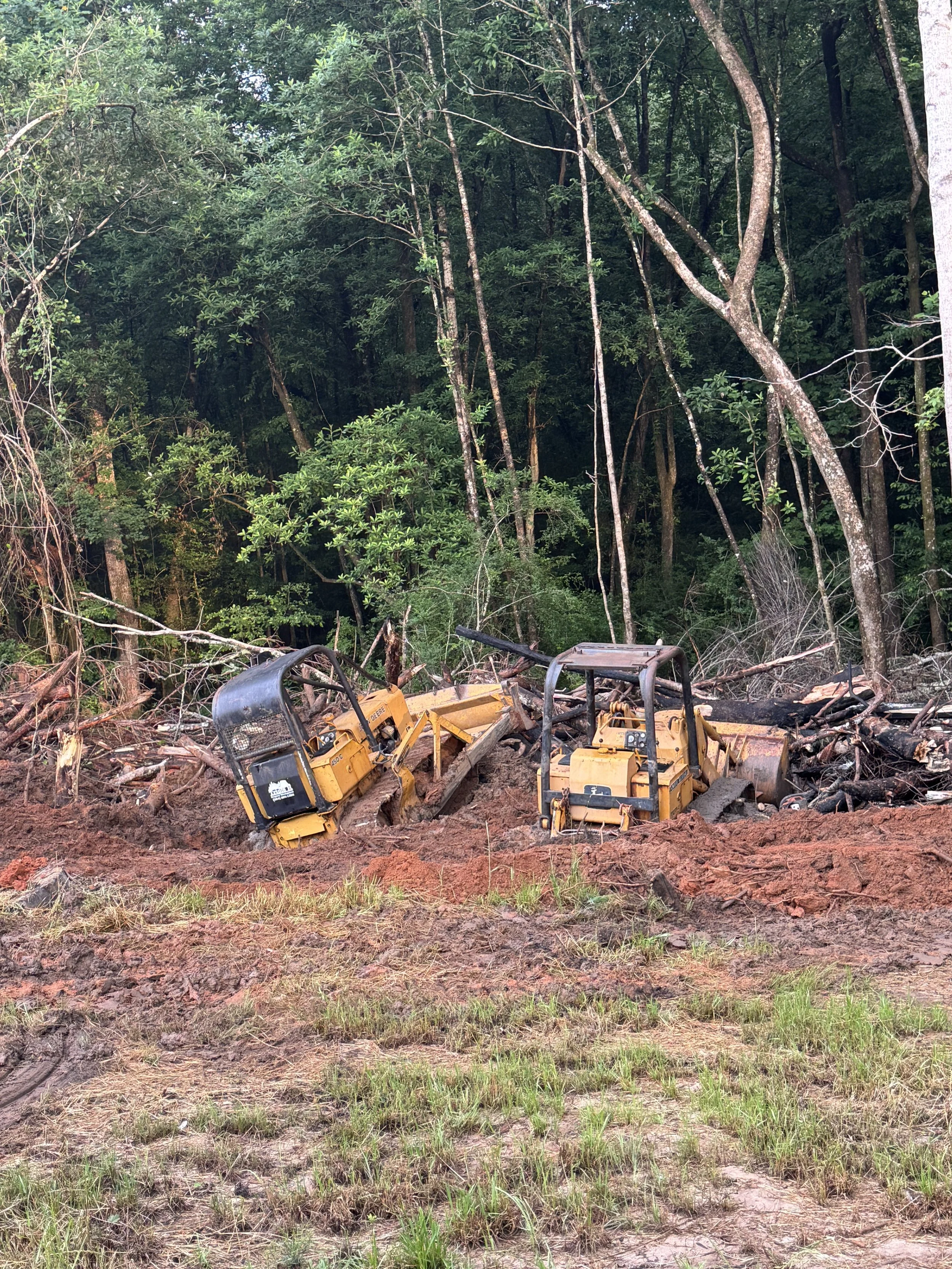 A cleared area of land with tree branches and roots, and yellow construction equipment in the foreground, surrounded by dense green forest.