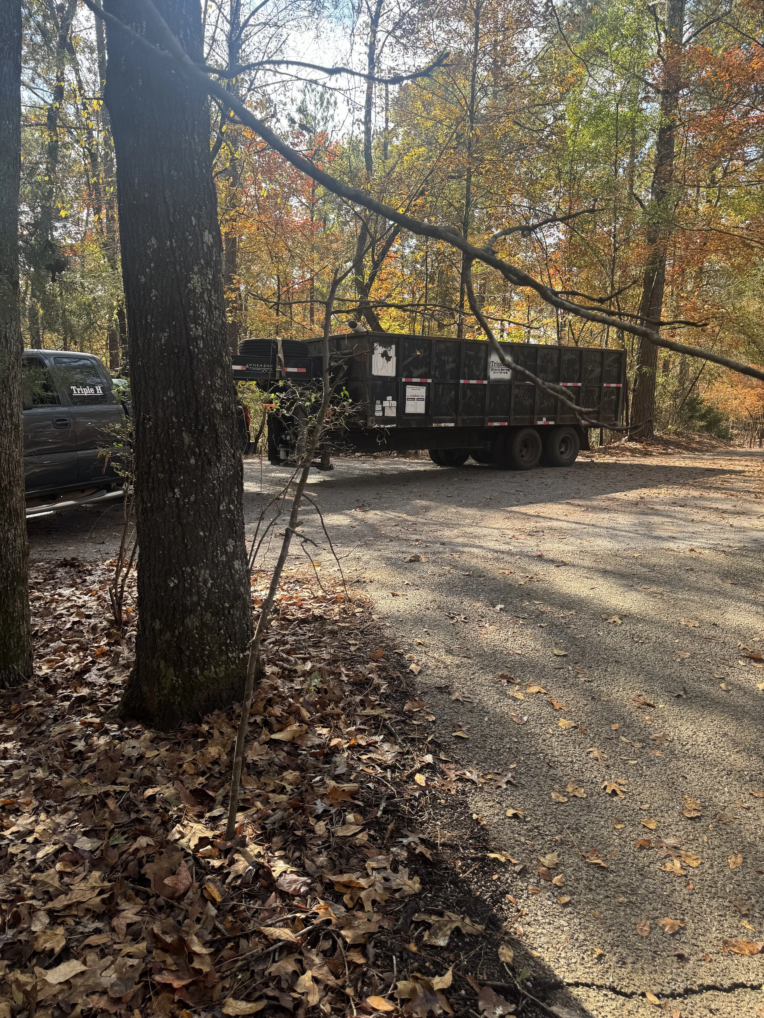 A parking area in a wooded area with trees and fallen leaves, featuring a black truck with a trailer attached and a dark-colored vehicle nearby.