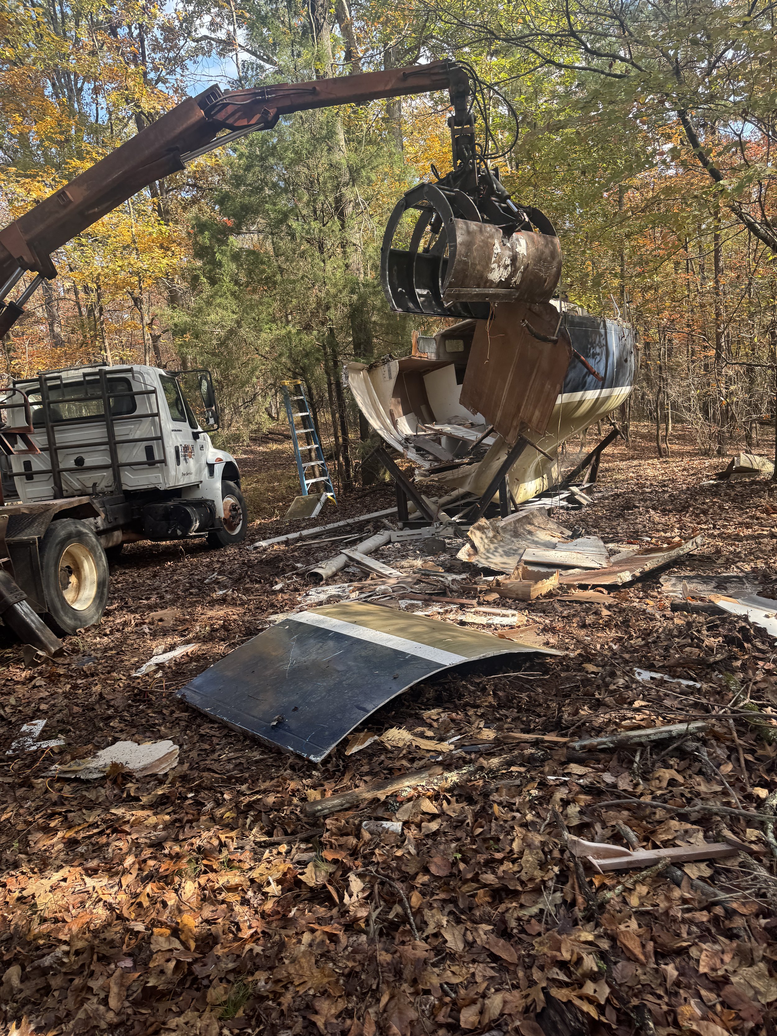Sail Boat being demoed by Triple H  being loaded into a grapple truck and ladder nearby, set in a wooded area with fall leaves on the ground.