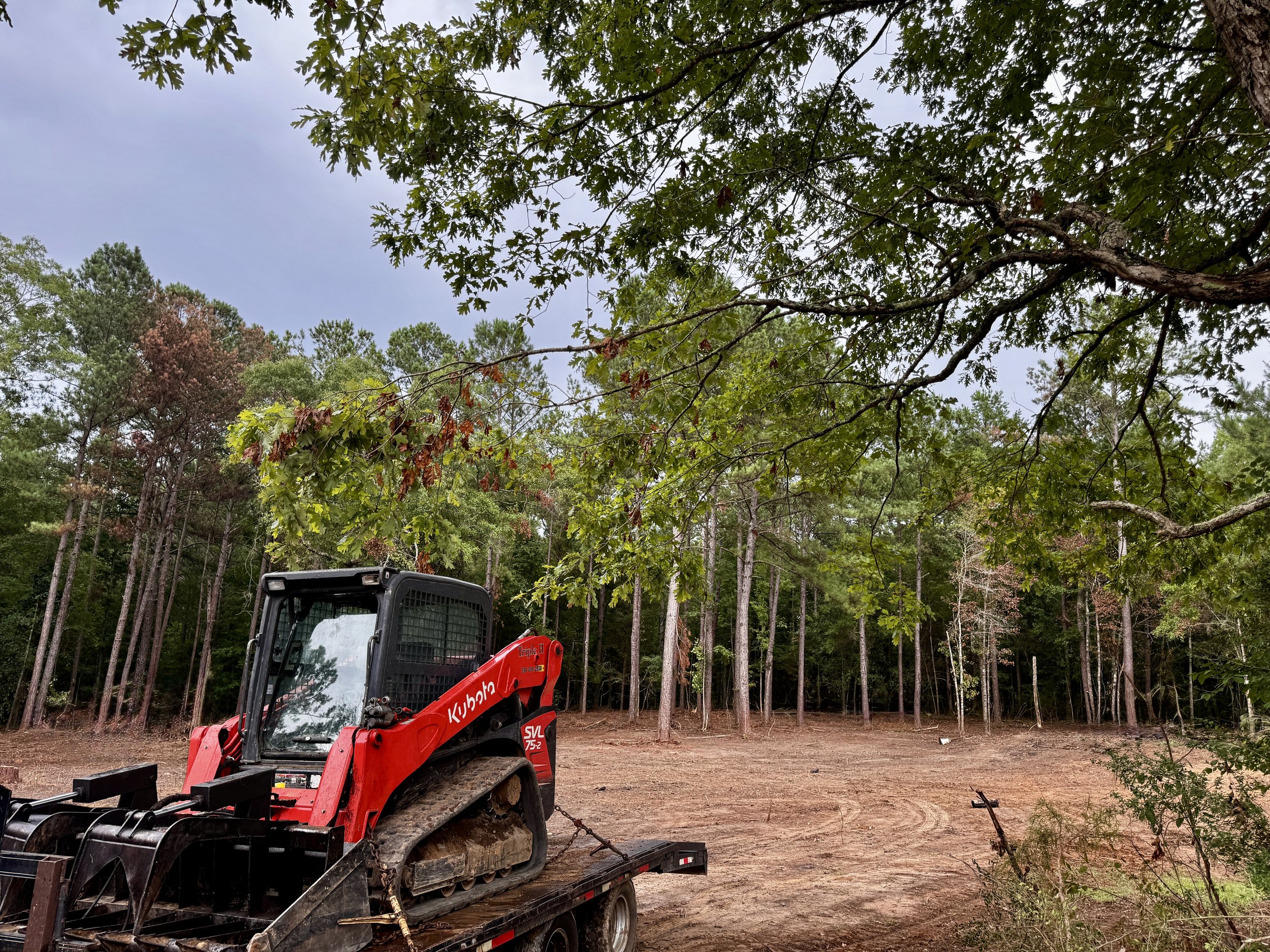 A small Kubota skid steer loader on a trailer in a cleared outdoor area with a forest of trees in the background.