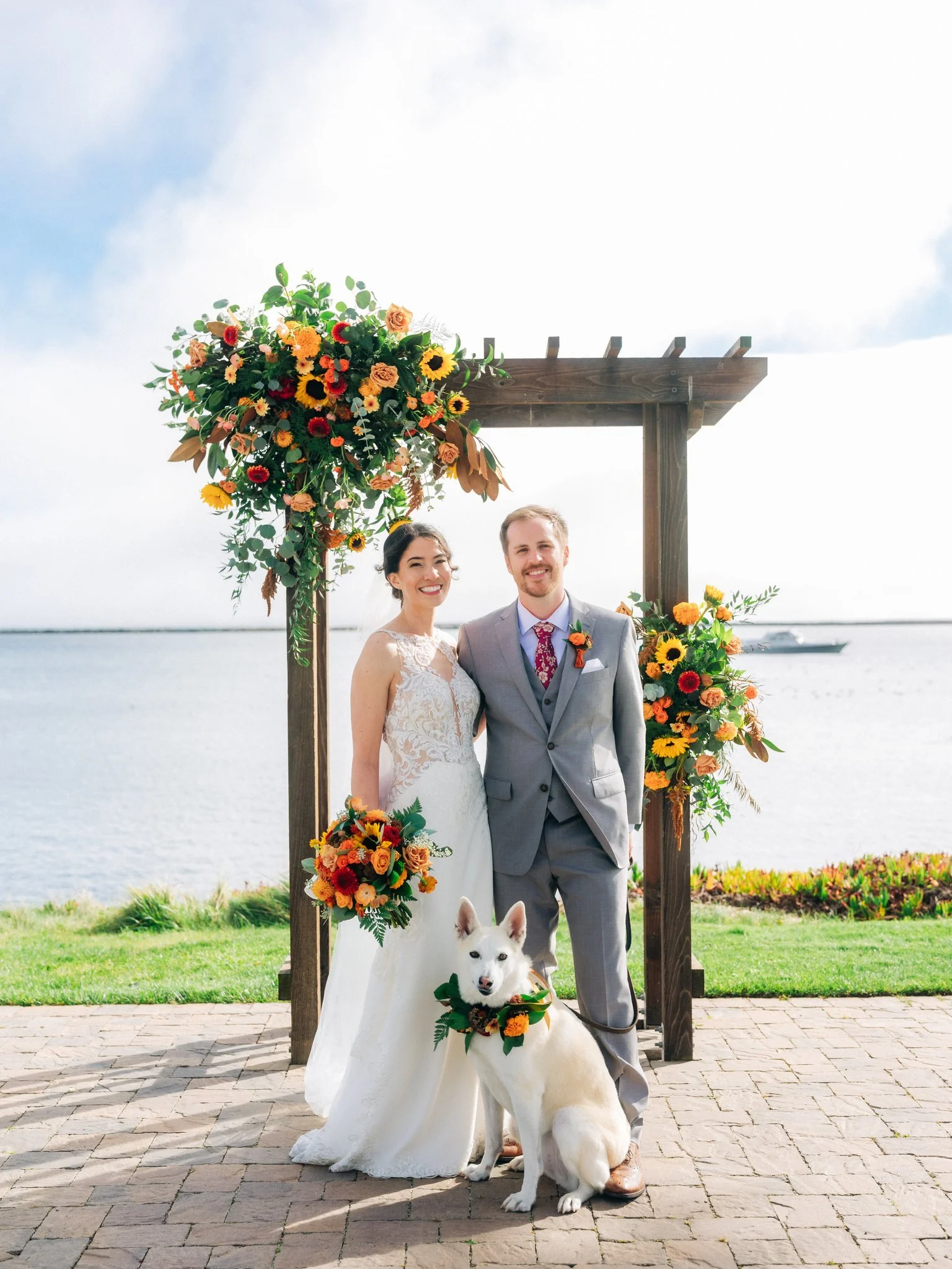 Image of the wedding arch. Backdrop of the ocean. Couple standing in front of wedding arch with fall flowers and their dog.