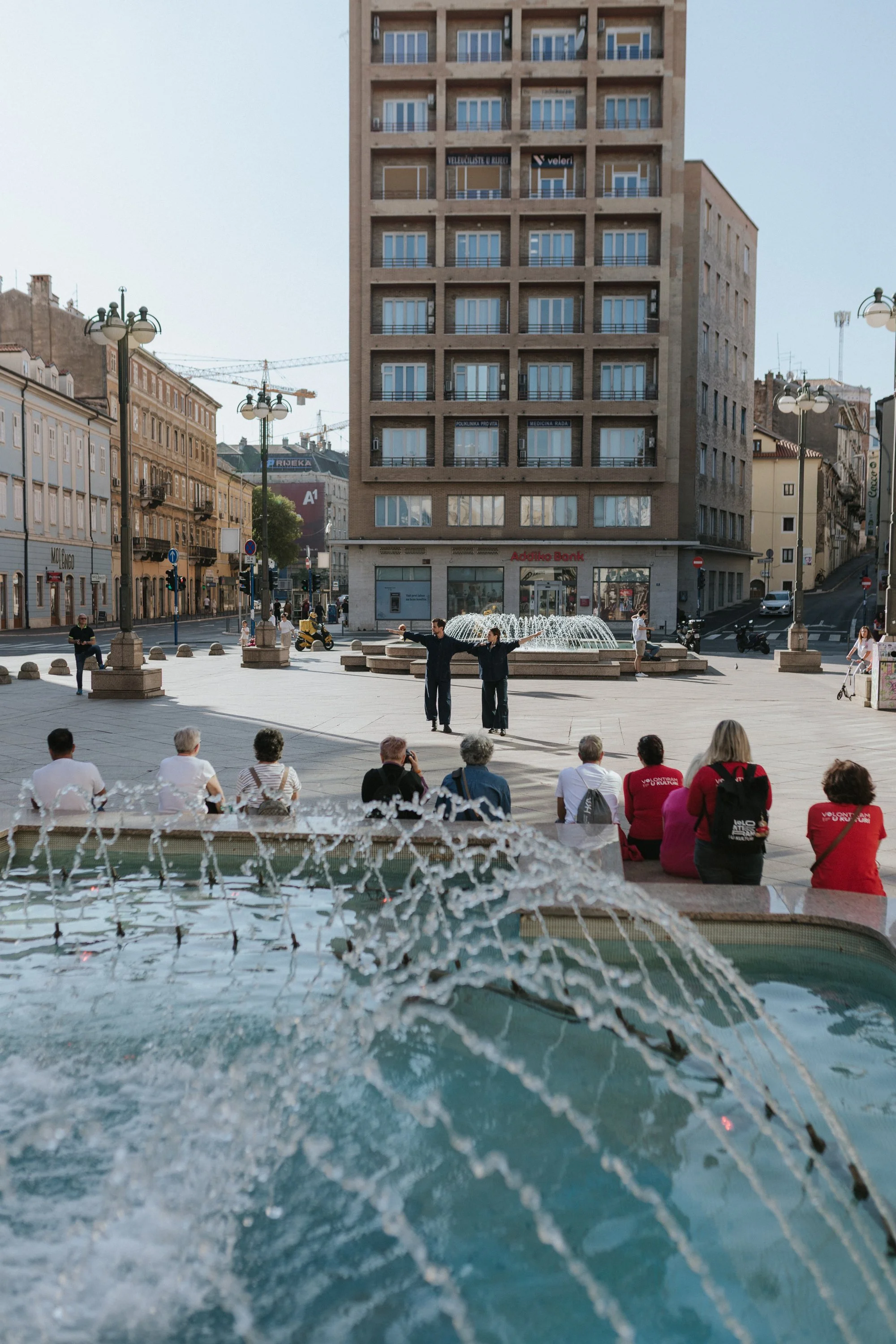 Seagulls Fly Early: the performers crossing a city square beside a fountain