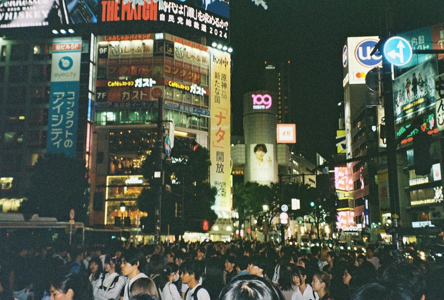 Night scene in a busy city intersection with numerous illuminated signs and billboards in Japanese, crowded with people waiting to cross.