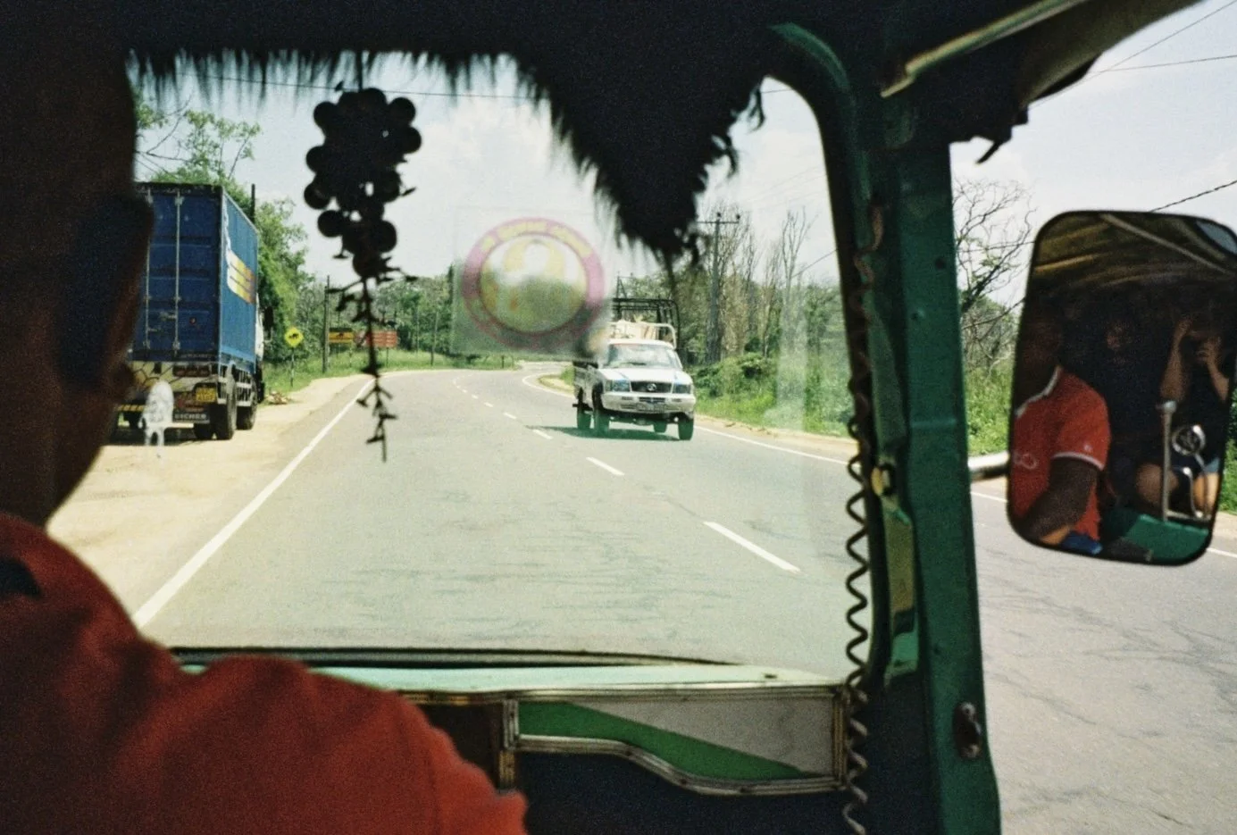 View from inside a vehicle on a winding rural road, showing a car approaching from the front and the side mirror with a person on the right. The scene includes trees and power lines along the roadside.