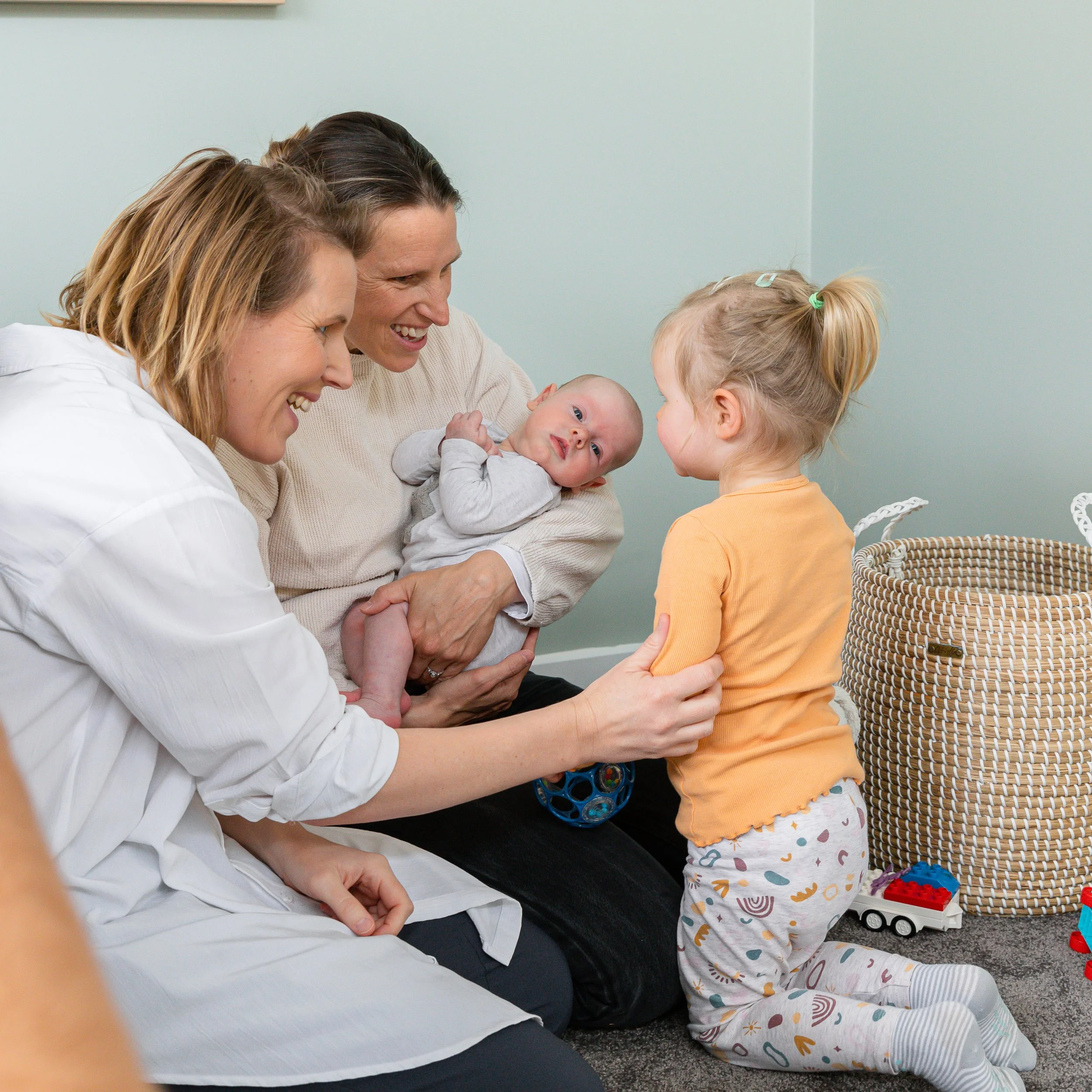 Three women and a young girl with a baby in her arms in a room, engaging in a friendly interaction. The women are smiling, and the girl appears to be talking to the baby. There are toys and a basket in the background.