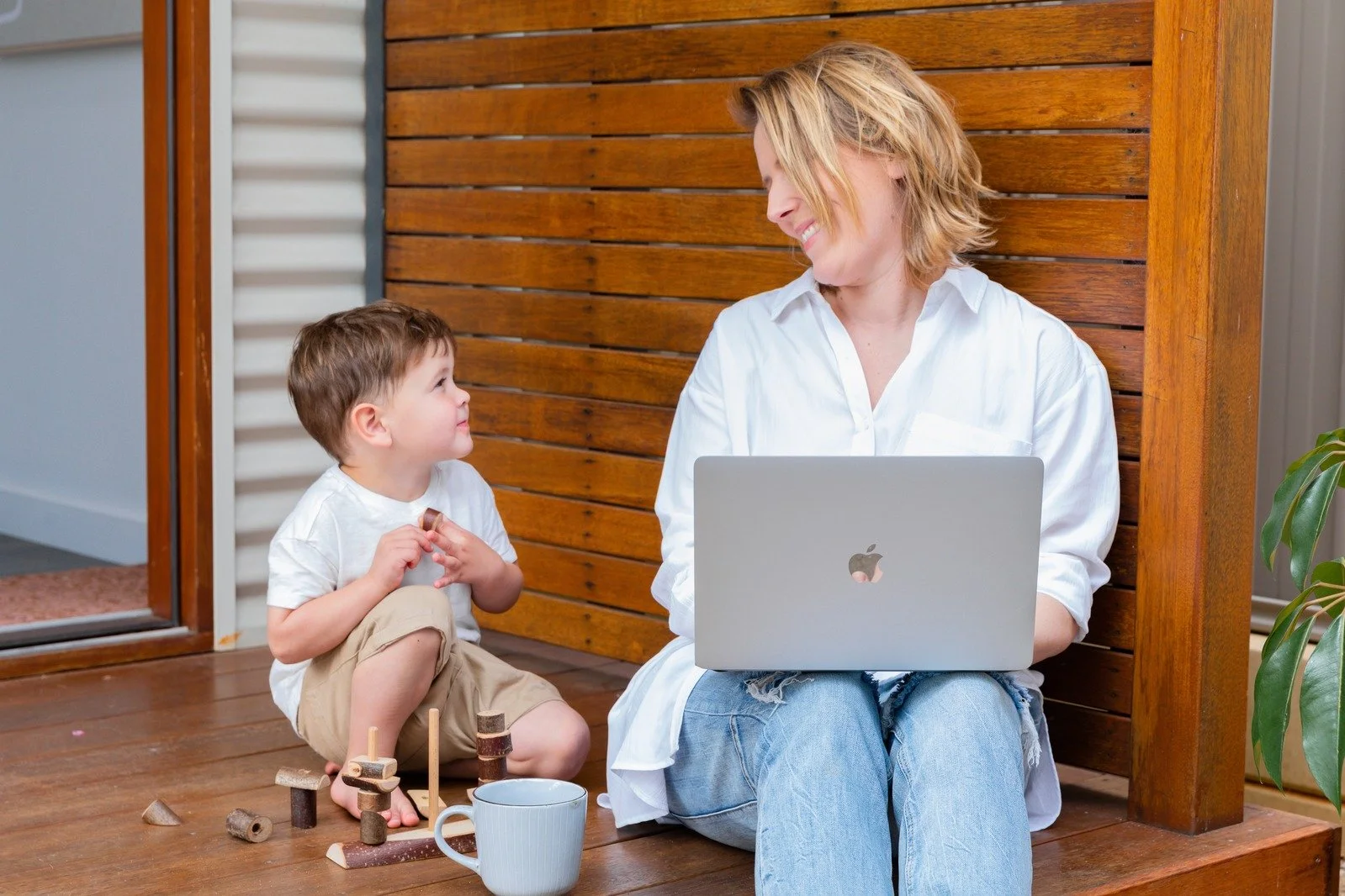 A woman and a young boy sitting on a wooden porch, with the woman using a laptop and the boy playing with wooden toys, engaging in a shared moment of happiness.