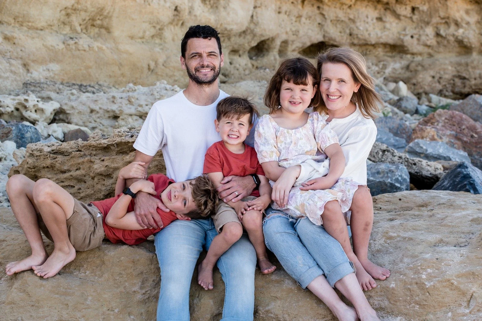 Family of six sitting on rocks at the beach, smiling and hugging each other.
