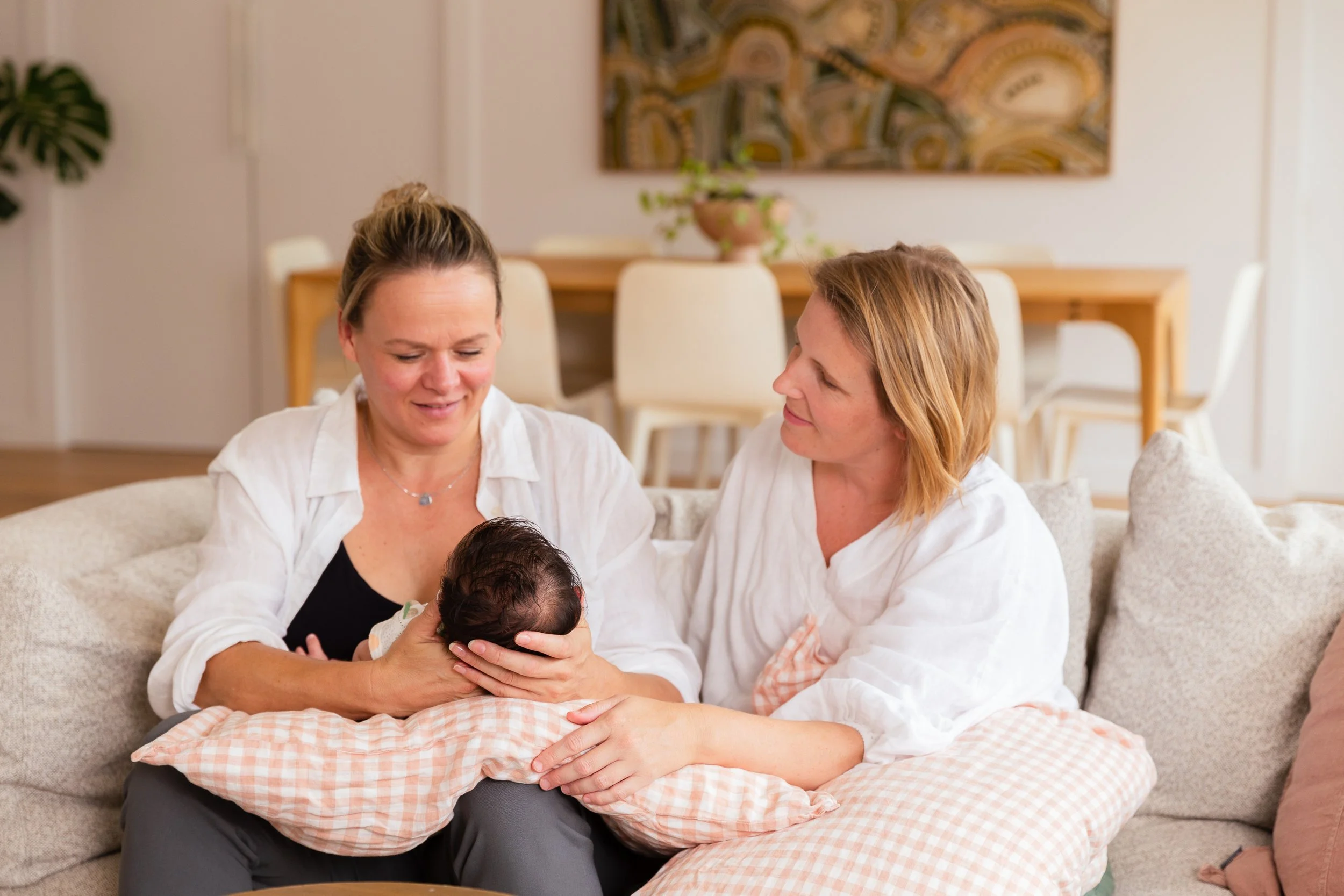 Two women, one holding a newborn baby, sitting on a sofa in a cozy living room, sharing a tender moment.