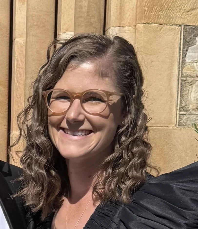 A woman with curly brown hair and glasses smiling outdoors against a tan stone wall.