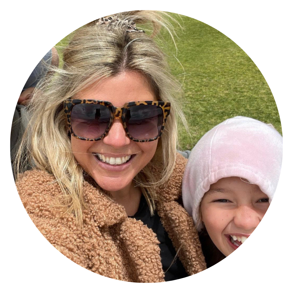A woman with blonde hair wearing large tortoise-shell sunglasses and a brown fuzzy jacket, smiling with a young girl who is wearing a pink hat, also smiling, outdoors on a grassy area.