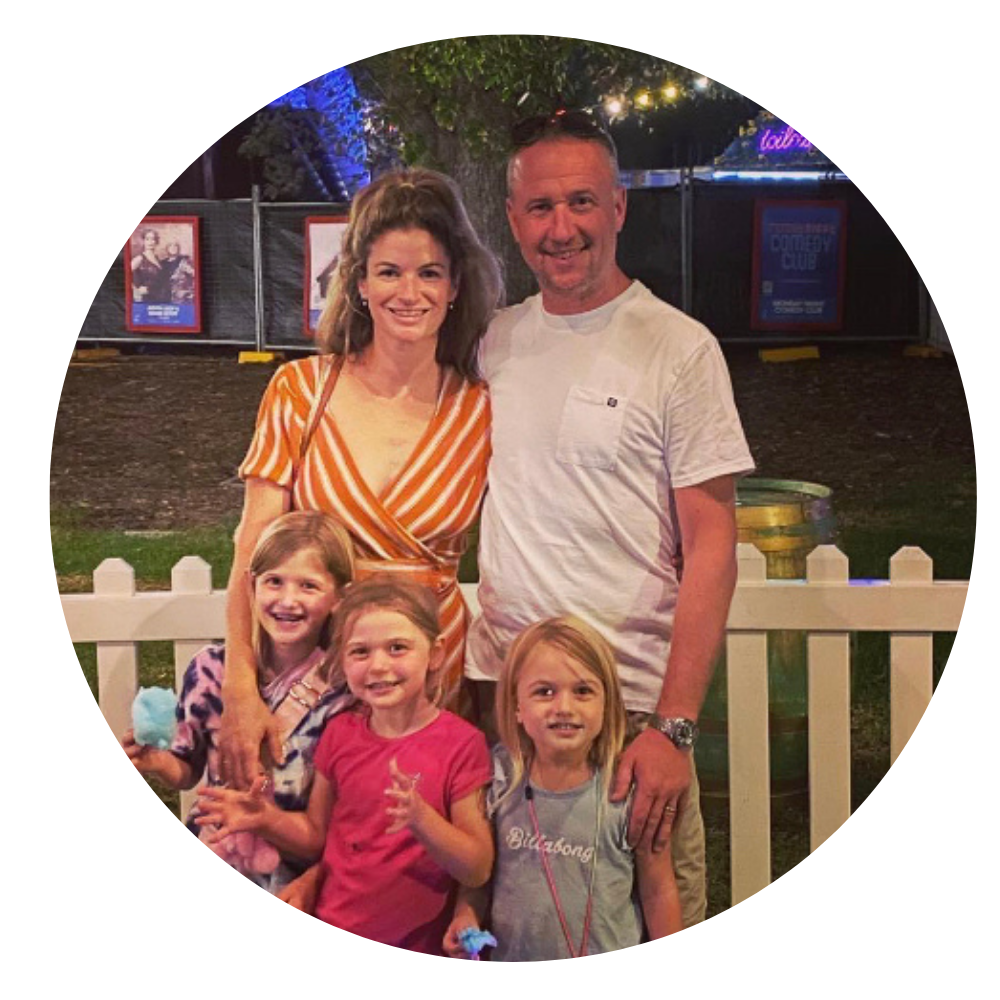 A family of five, including two adult women and three young girls, enjoying an outdoor event at night. They are smiling and posing in front of a white picket fence with colorful signs and banners in the background.