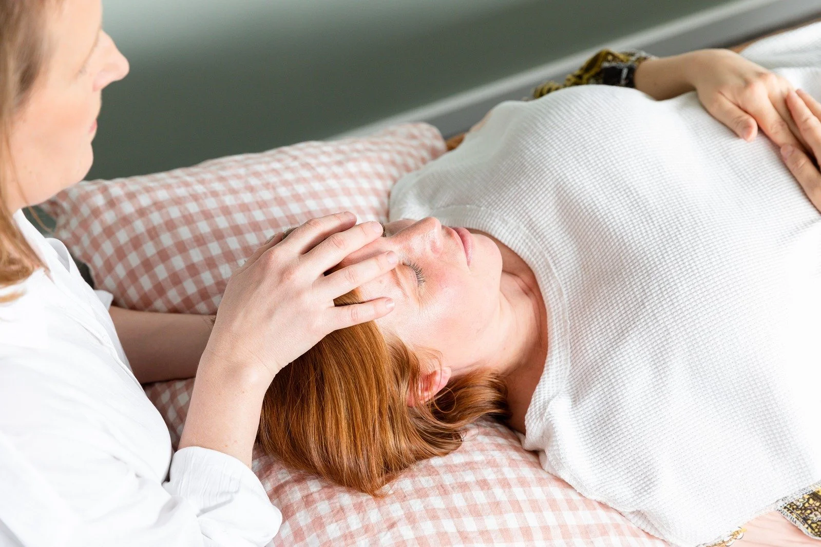 A woman lying in bed with her eyes closed, receiving a head massage from another woman who is gently holding her forehead. The woman on the bed has red hair and is wearing a white shirt, while the woman giving the massage has brown hair and is wearing a white shirt.