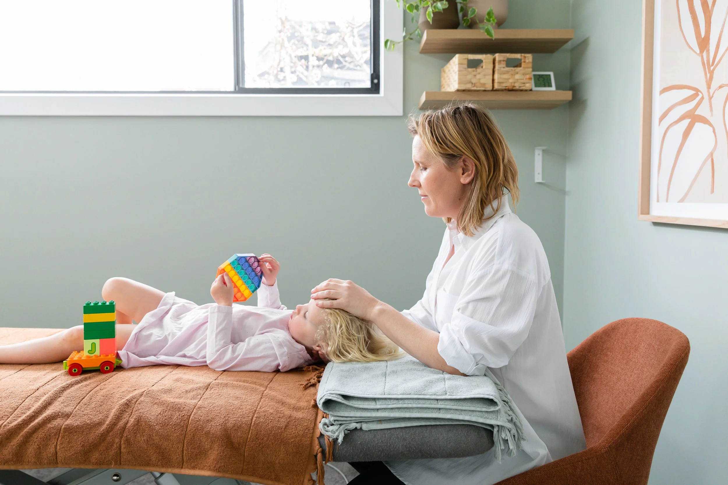 A doctor or therapist examining a young girl lying on a bed, with a stacking rainbow toy in her hands. The girl is relaxed and the setting appears to be a pediatric clinic or therapy room, with a window, shelves with decor, and artwork on the wall.