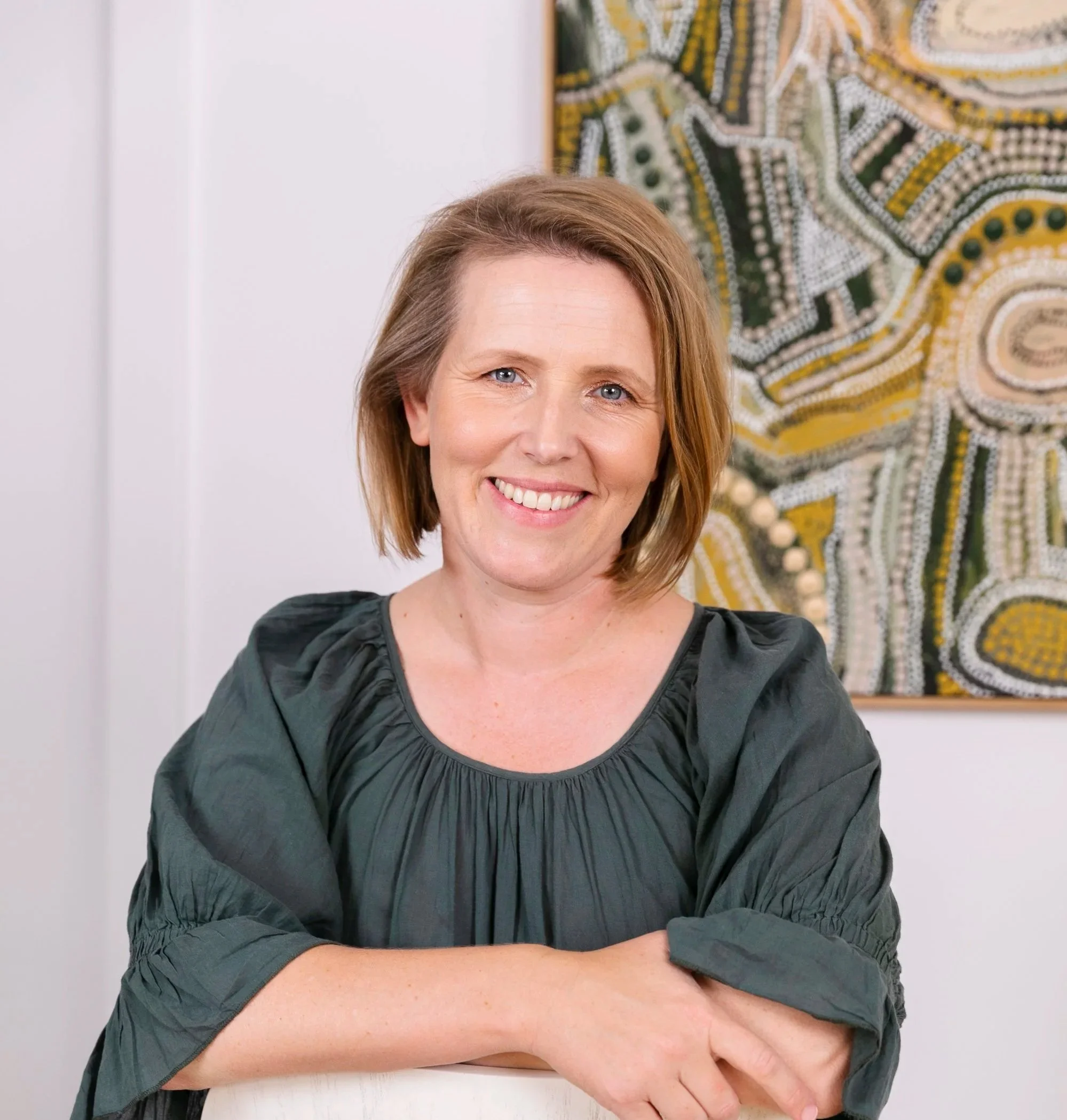 A woman with short brown hair and a smile, sitting in front of a colorful abstract painting in a bright room.