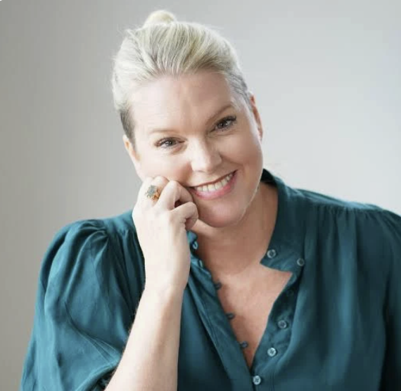 A woman with blonde hair styled in a bun, smiling and resting her chin on her hand, wearing a teal blouse.