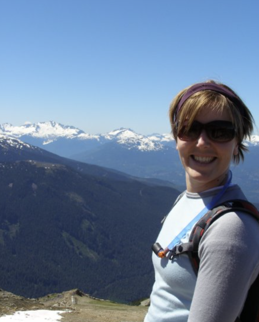 Smiling person wearing sunglasses and a backpack hiking in mountainous area with snow-capped peaks in the background.