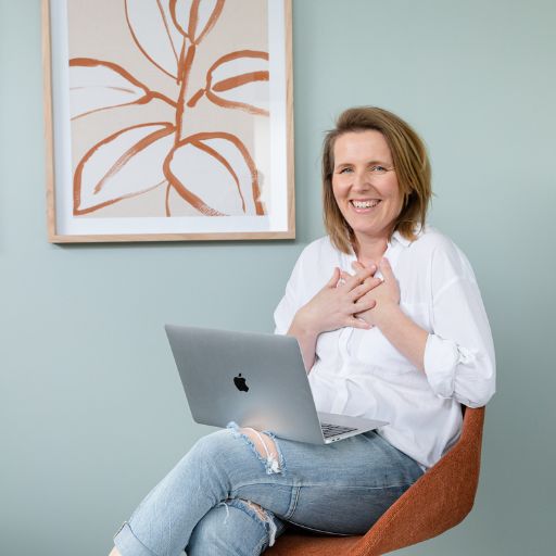A woman smiling happily while sitting on an orange chair with a silver laptop on her lap, in a room with light blue walls and a large abstract artwork of a leaf design on the wall.