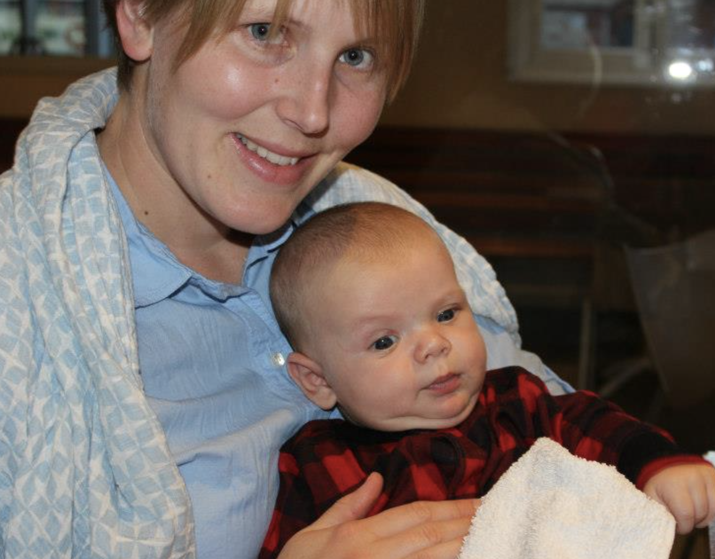 A woman with short light hair, wearing a light blue shirt and a patterned shawl, smiling and holding a young baby with short light hair, wearing a red and black plaid shirt, wrapped in a towel, in an indoor setting.