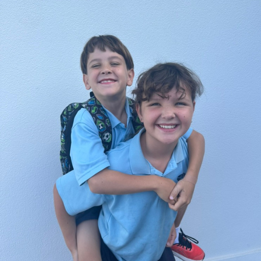 Two young boys smiling and playing together against a white wall. One boy is giving the other a piggyback ride.